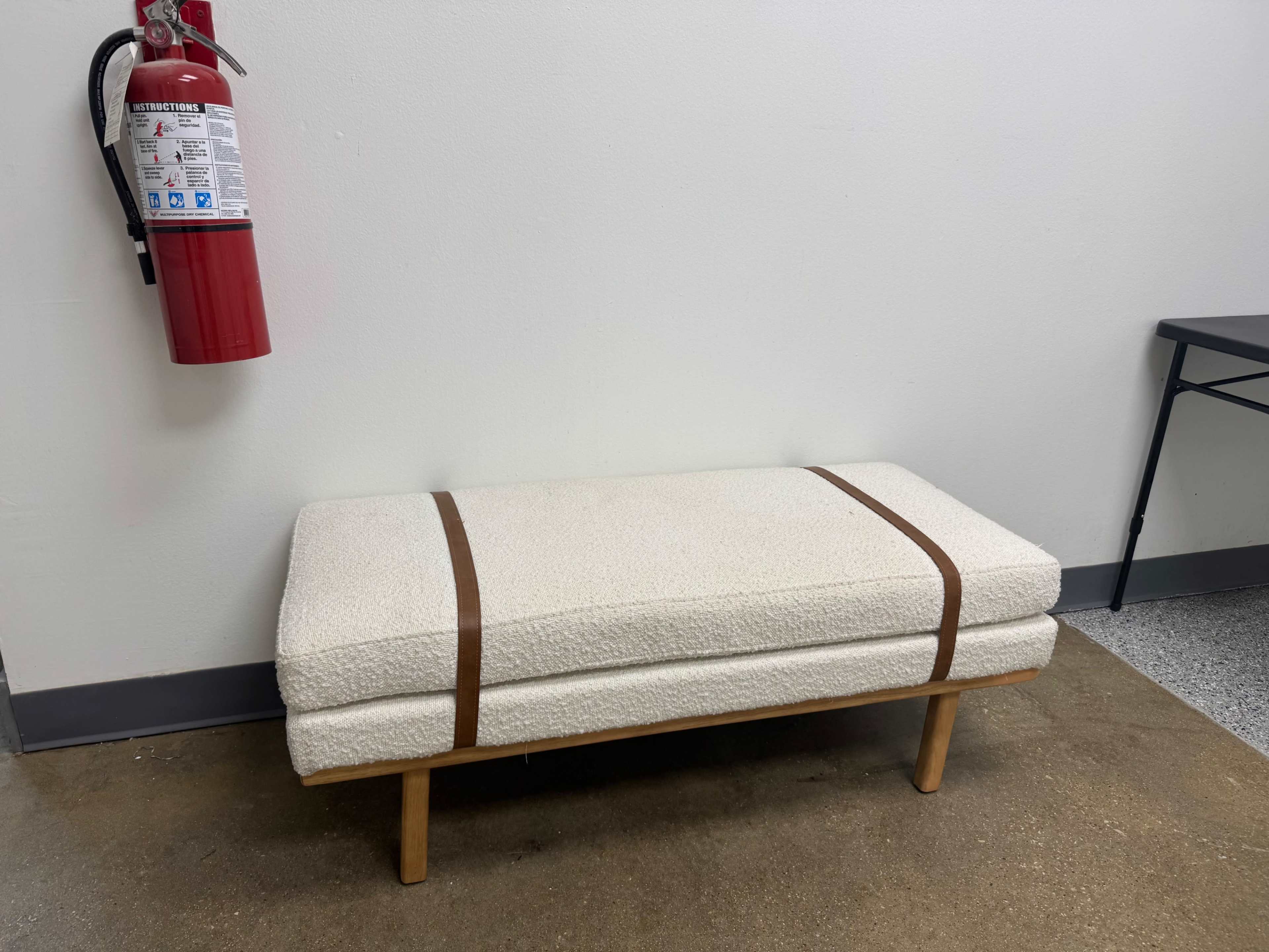 A cushioned bench with a wooden frame is placed against a wall next to a fire extinguisher.