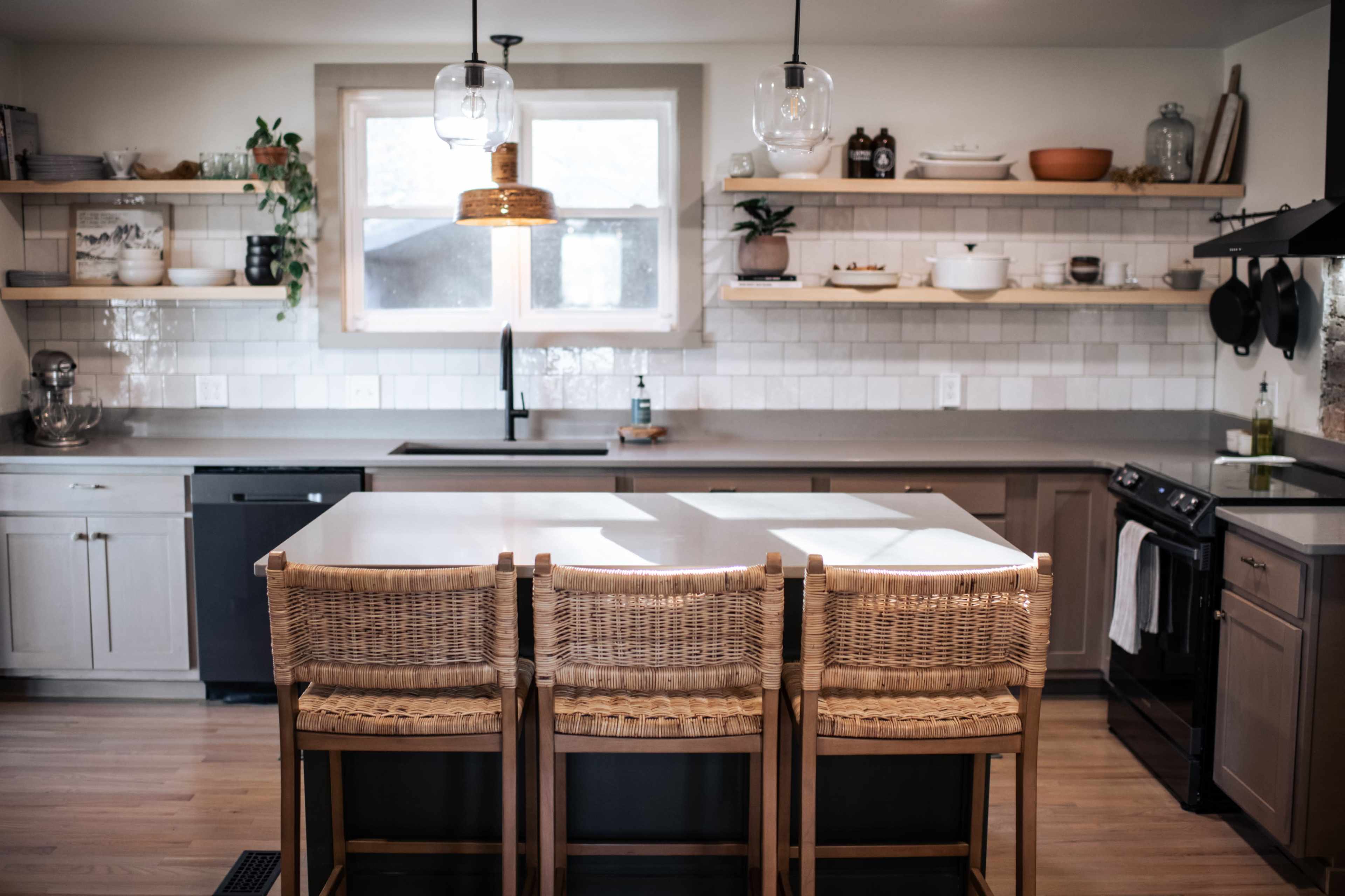 A modern kitchen features a large island with three woven bar stools, open shelving displaying kitchenware, and a window above the sink.