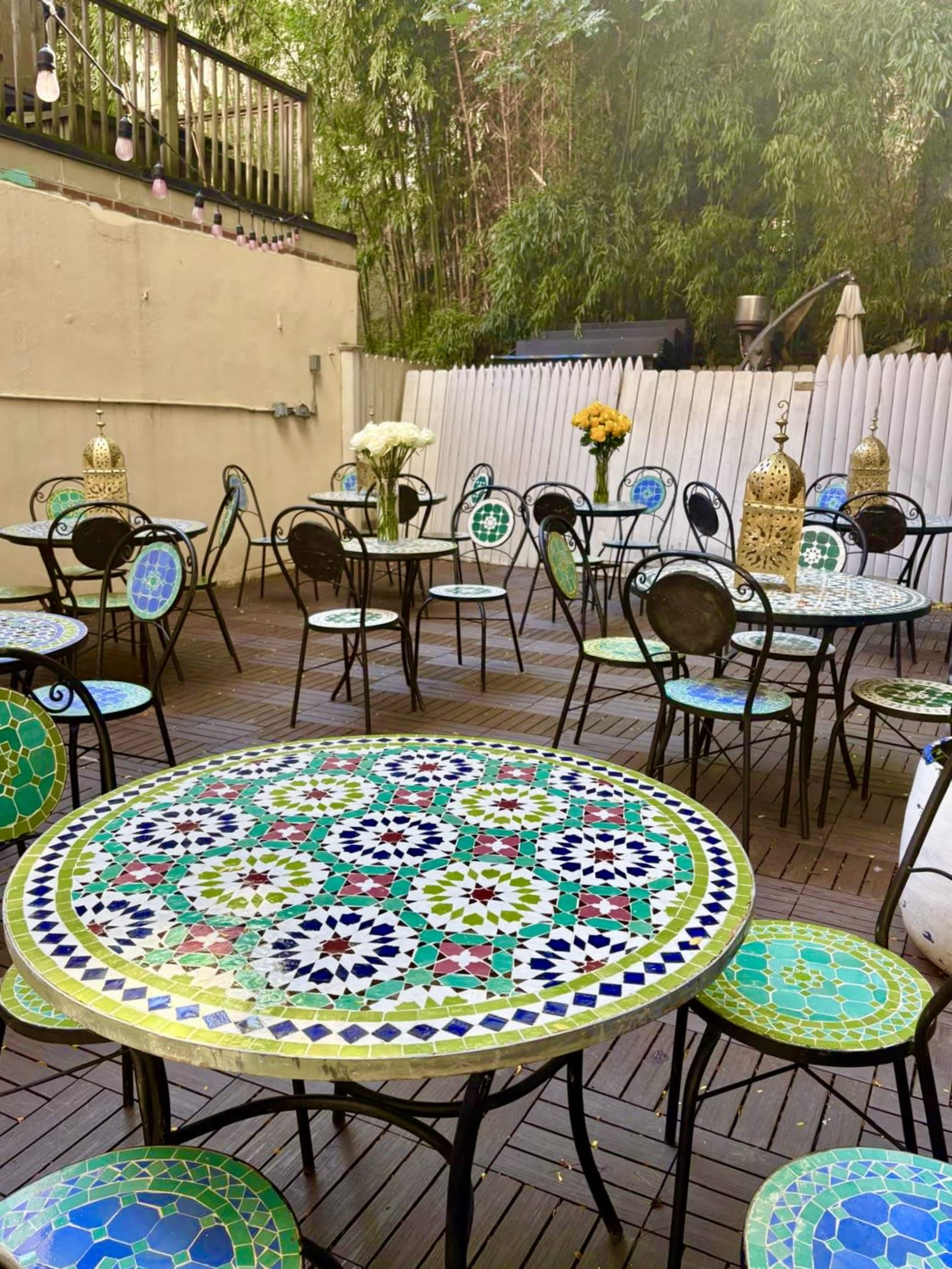 The scene features a courtyard with several mosaic-topped tables and wrought-iron chairs arranged on a wooden deck, surrounded by greenery and decorative elements.