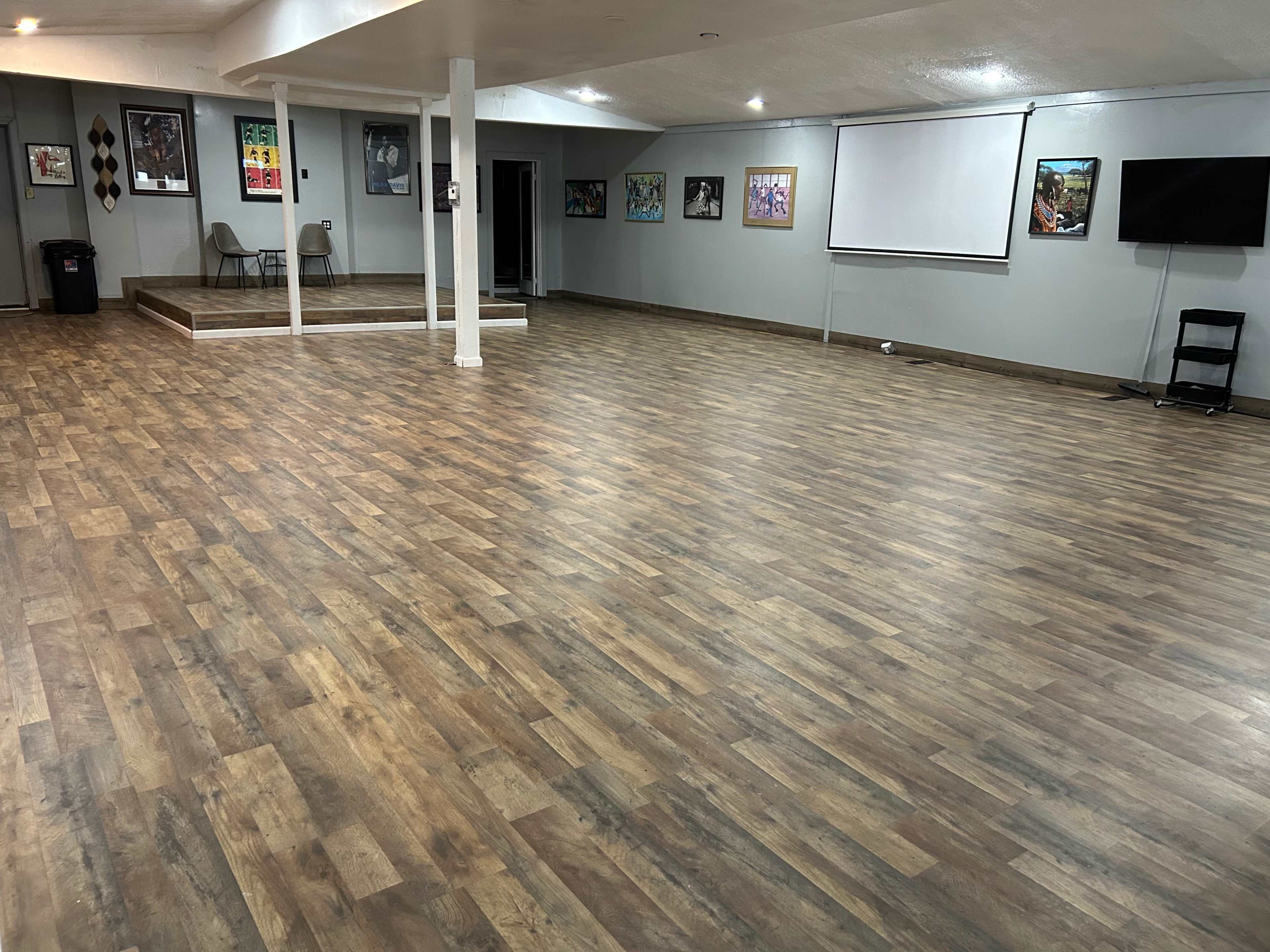 An empty, well-lit room with wooden flooring, a small stage area, and several framed artworks on the walls.