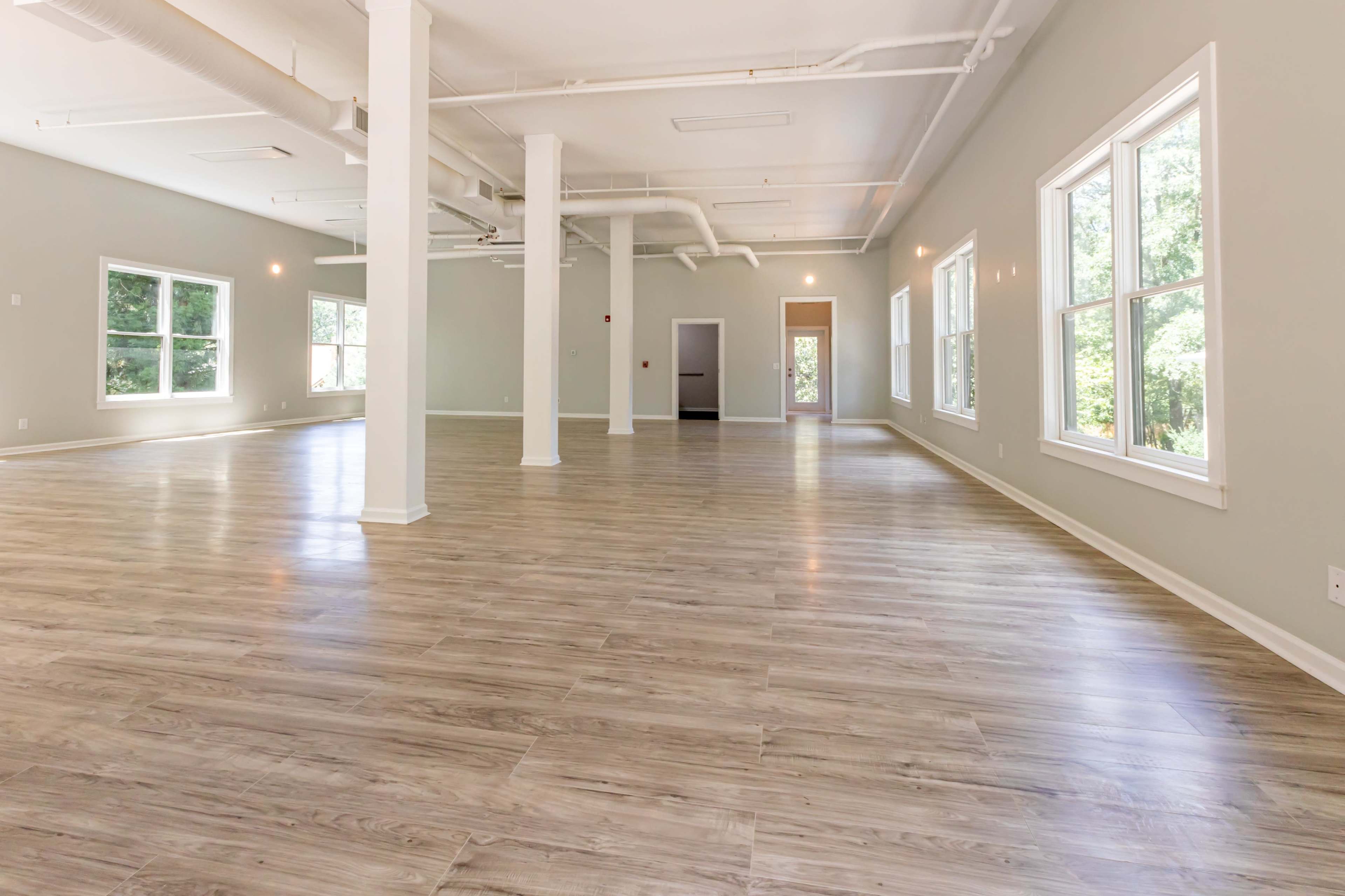 The image depicts a spacious, empty interior room with light-colored walls, large windows, and wooden flooring.