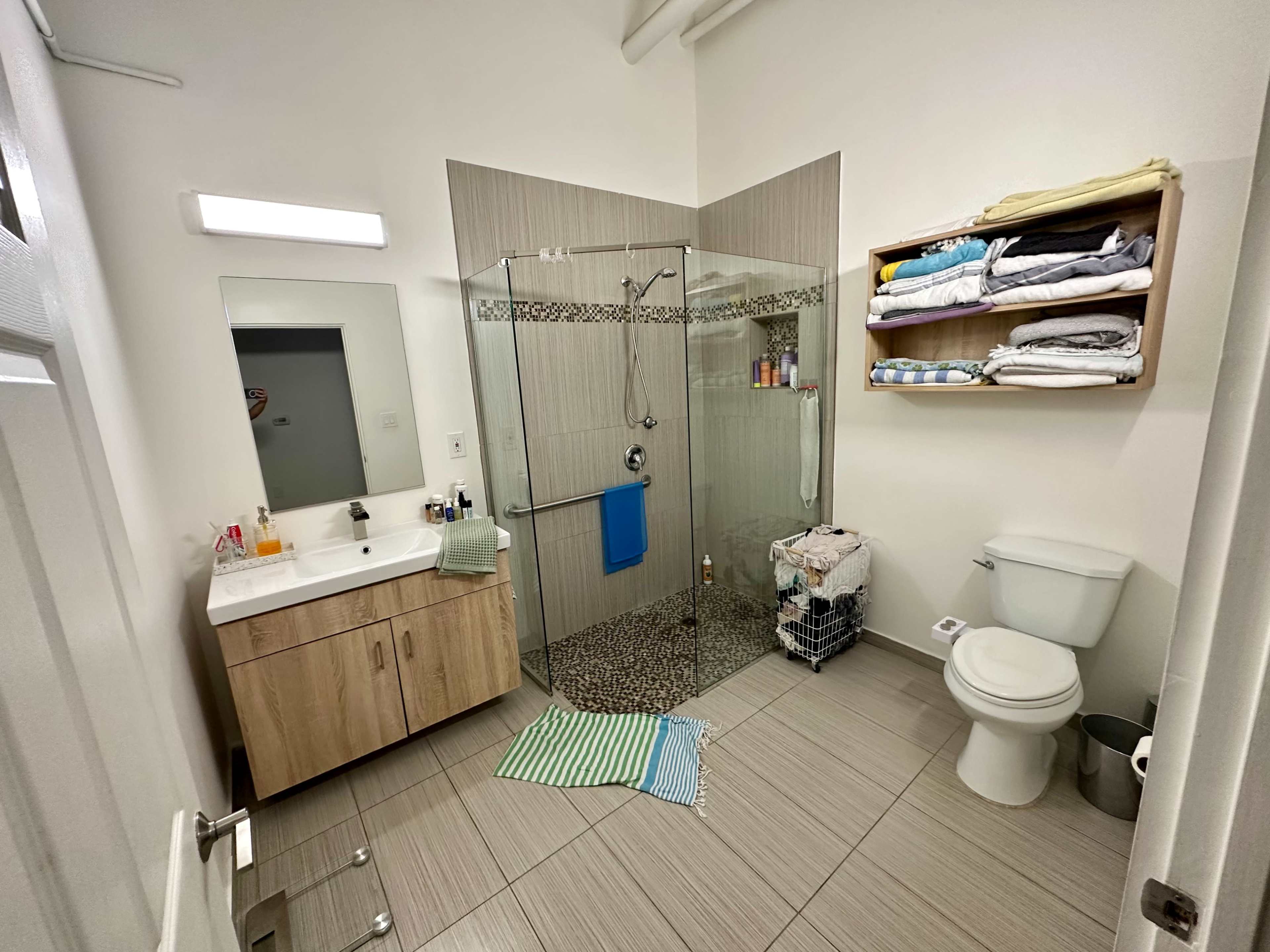 The image shows a modern bathroom featuring a glass shower, a wooden vanity with a mirror, a toilet, and shelves with neatly stacked towels.