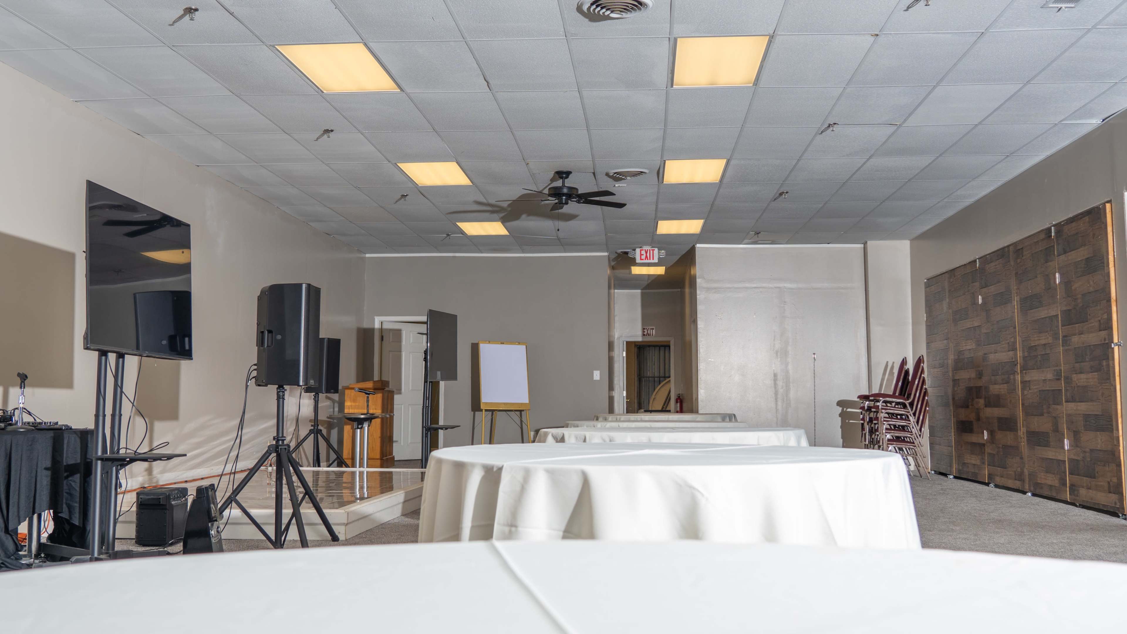 A spacious indoor event room featuring round tables covered with white tablecloths, and equipment set up for a presentation or meeting.