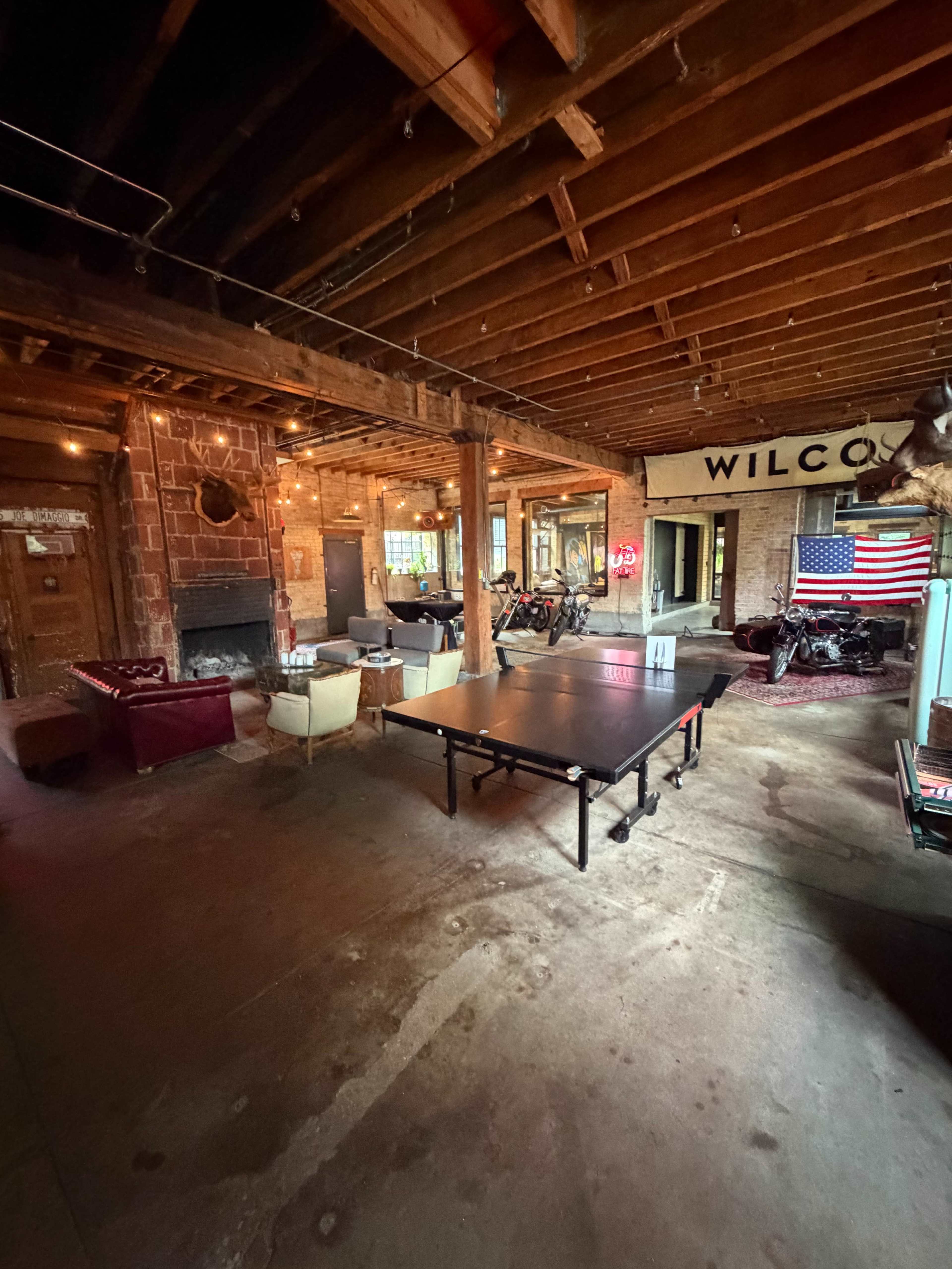 The image shows an indoor recreational space featuring a ping pong table, several seating areas, a fireplace, and displayed bicycles with an American flag on the wall.