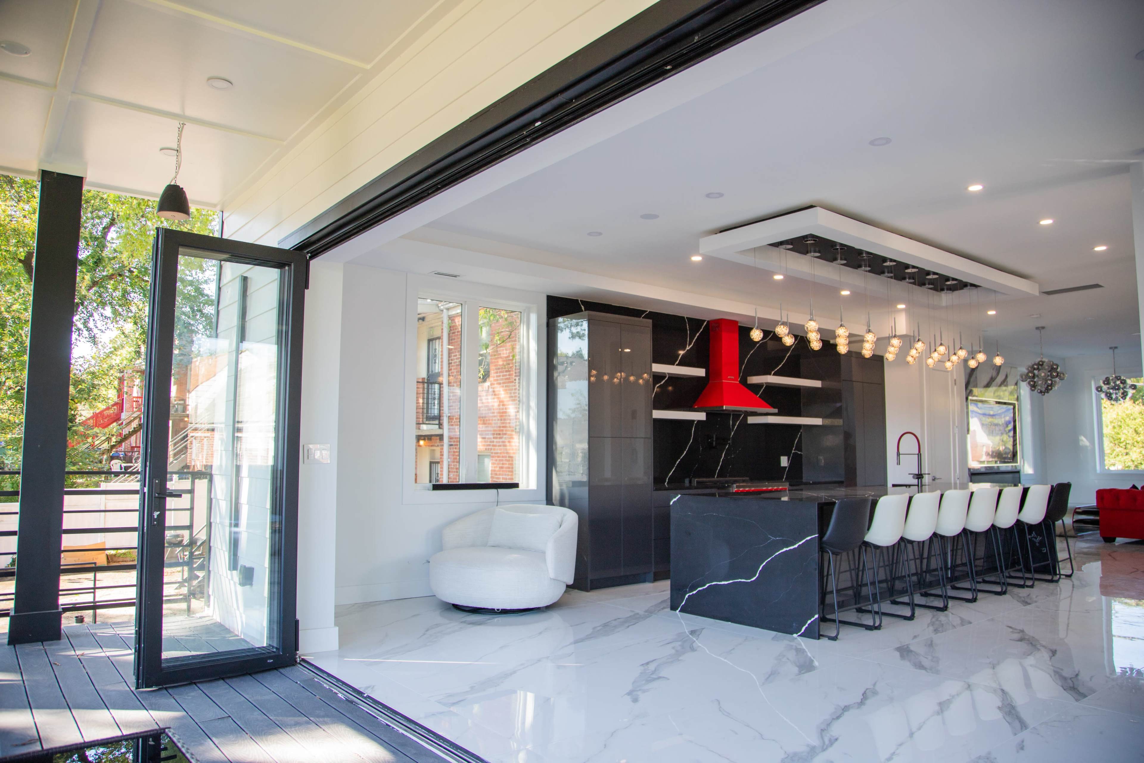 The image shows a modern kitchen with a large central island, sleek cabinetry, and an open layout that connects to an outdoor space through large glass doors.
