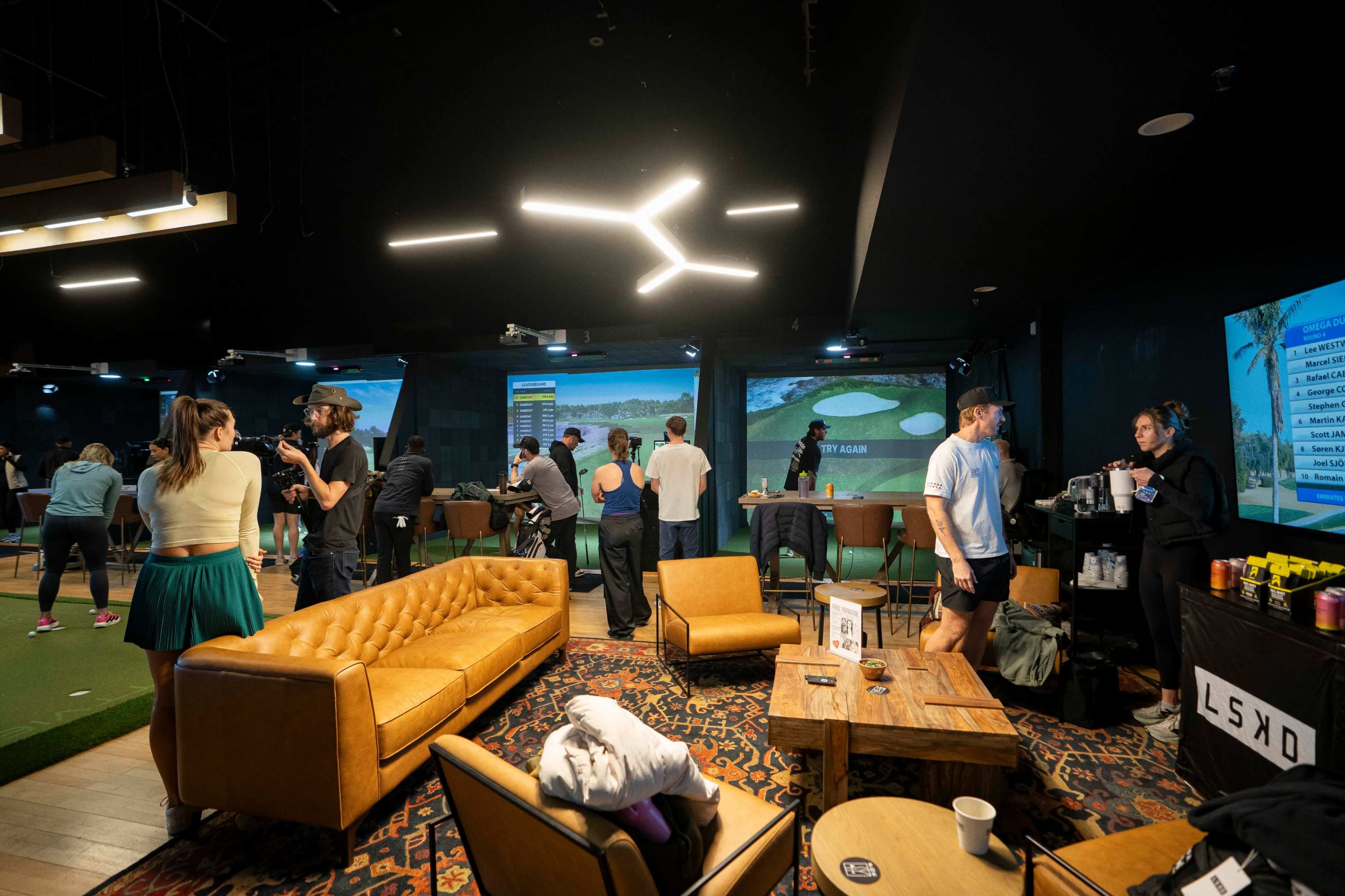 The image shows a social indoor golfing venue with people engaging in activities, surrounded by golf simulators and casual seating arrangements.