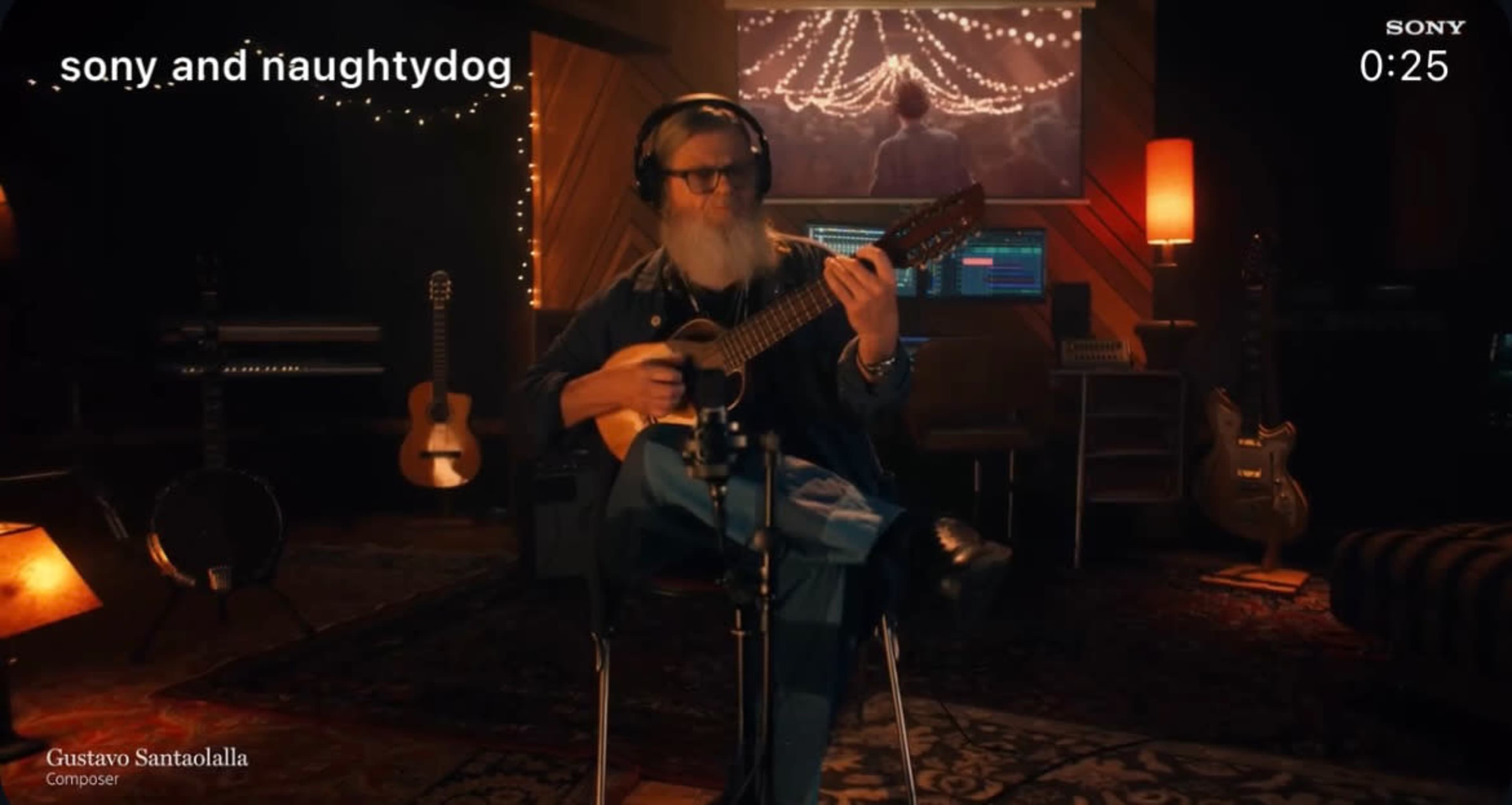 A person with a long beard plays a guitar in a warmly lit music studio surrounded by various musical instruments.