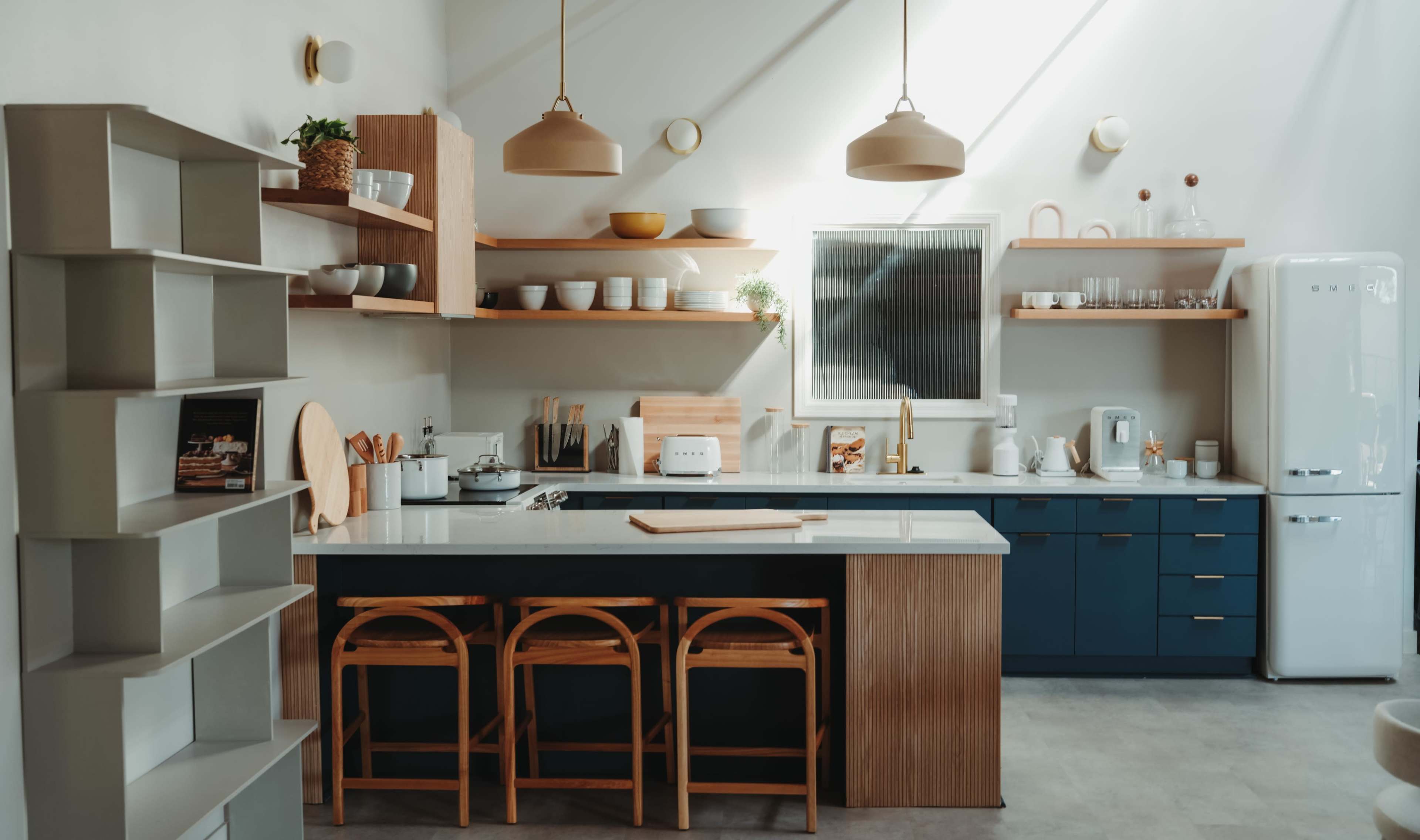 A modern kitchen features a blue and wooden color scheme with open shelving, pendant lights, and bar stools around an island.