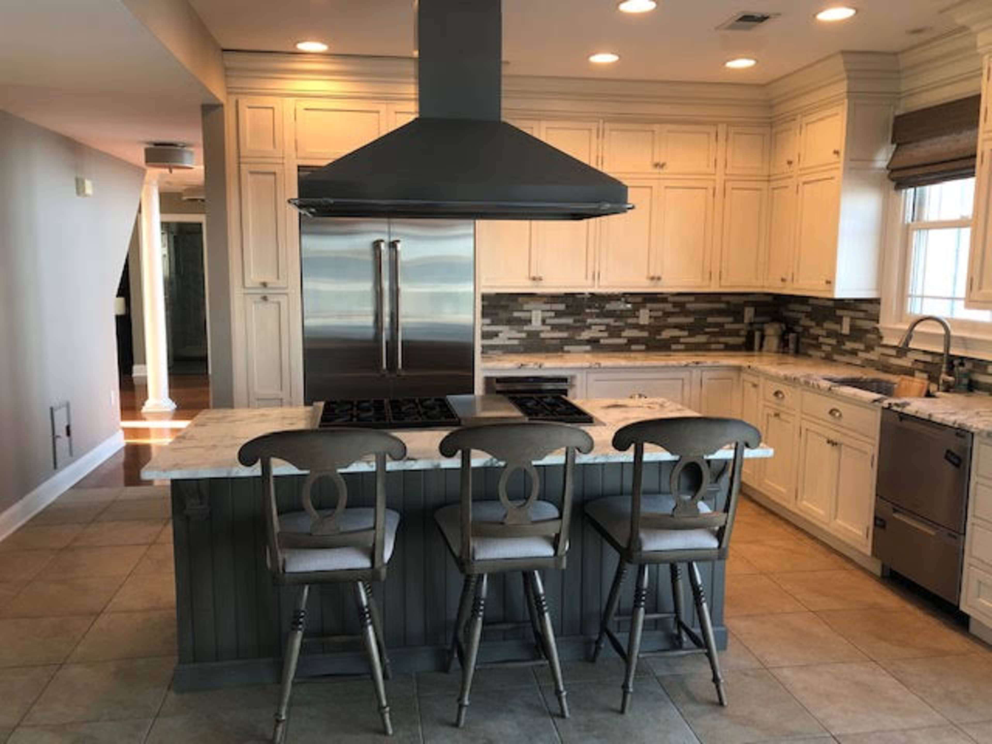 A modern kitchen with a central island, four bar stools, and white cabinetry, featuring a stainless steel range hood and backsplash of varied tiles.