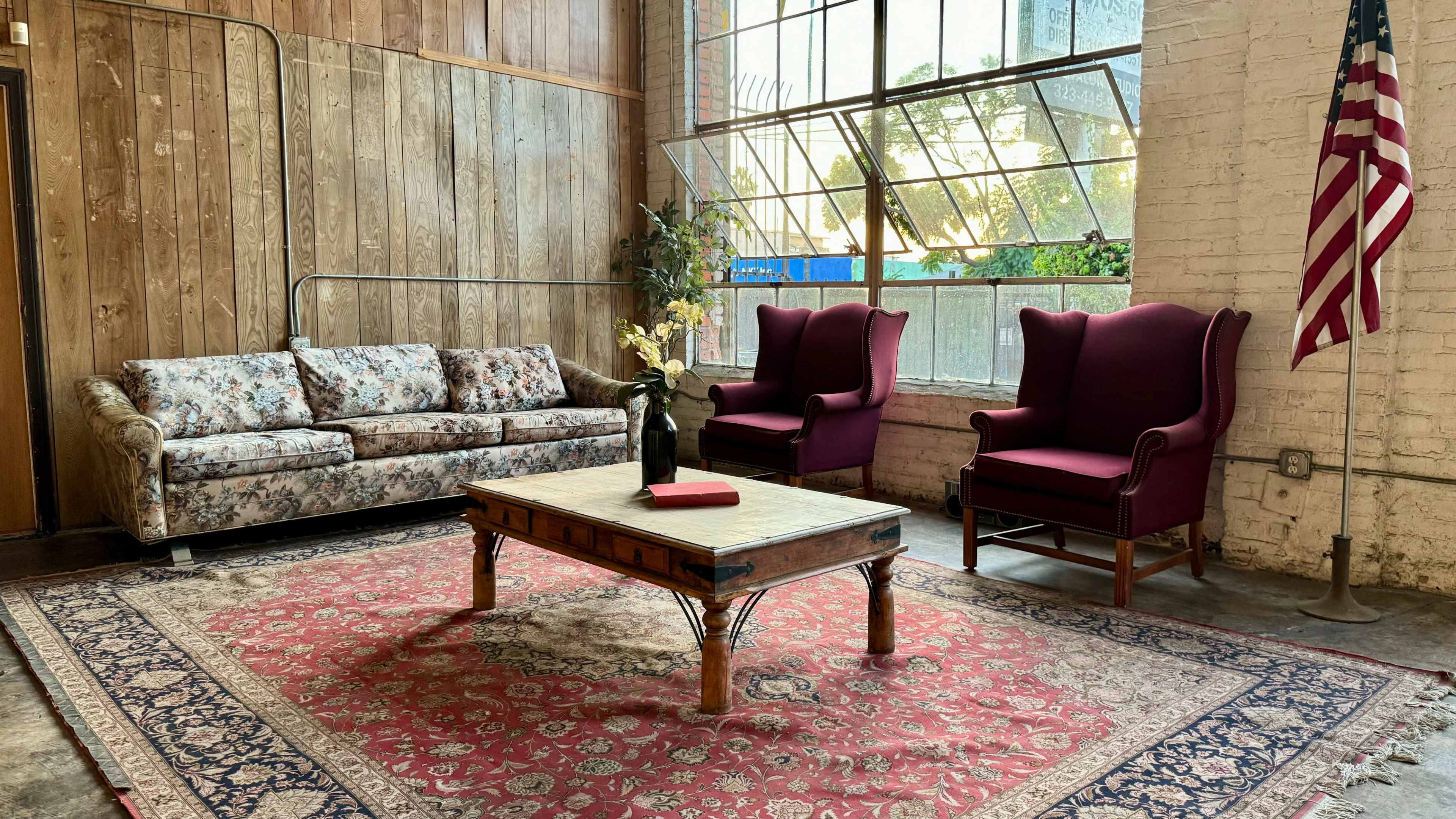 The image shows a cozy sitting area featuring a floral couch, two burgundy armchairs, and a wooden coffee table on a patterned rug, with large windows letting in natural light.