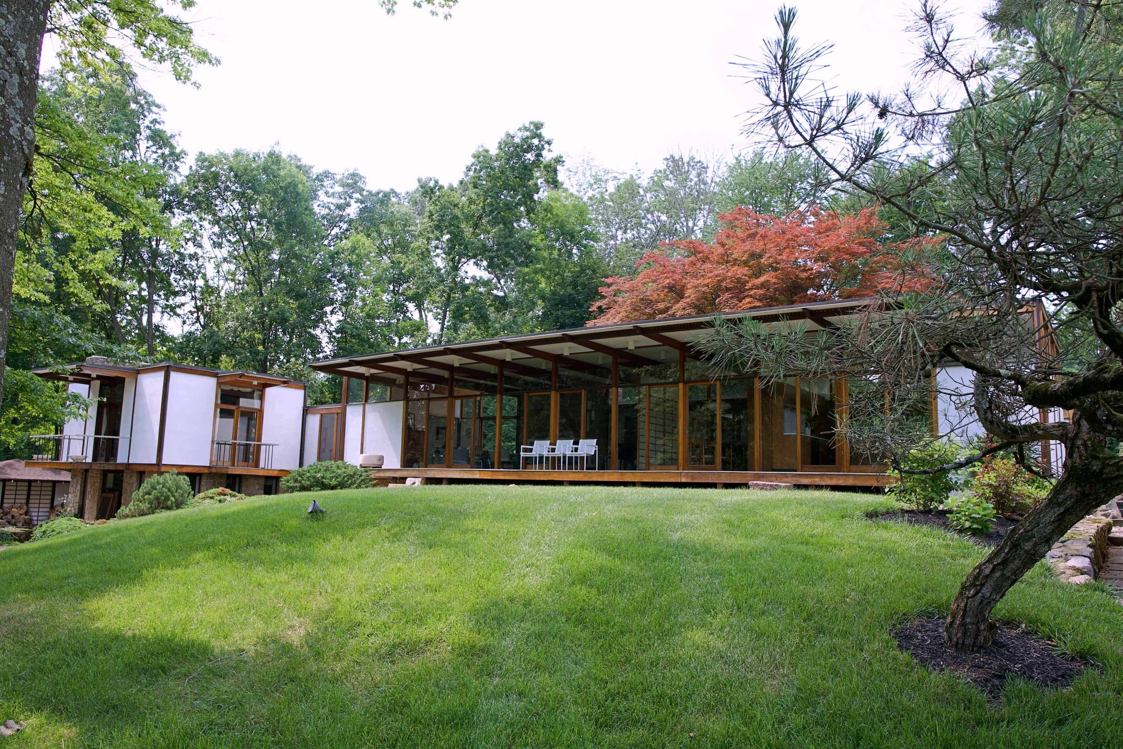 A modern, two-section house with large windows set on a grassy slope, surrounded by trees and landscaped greenery.