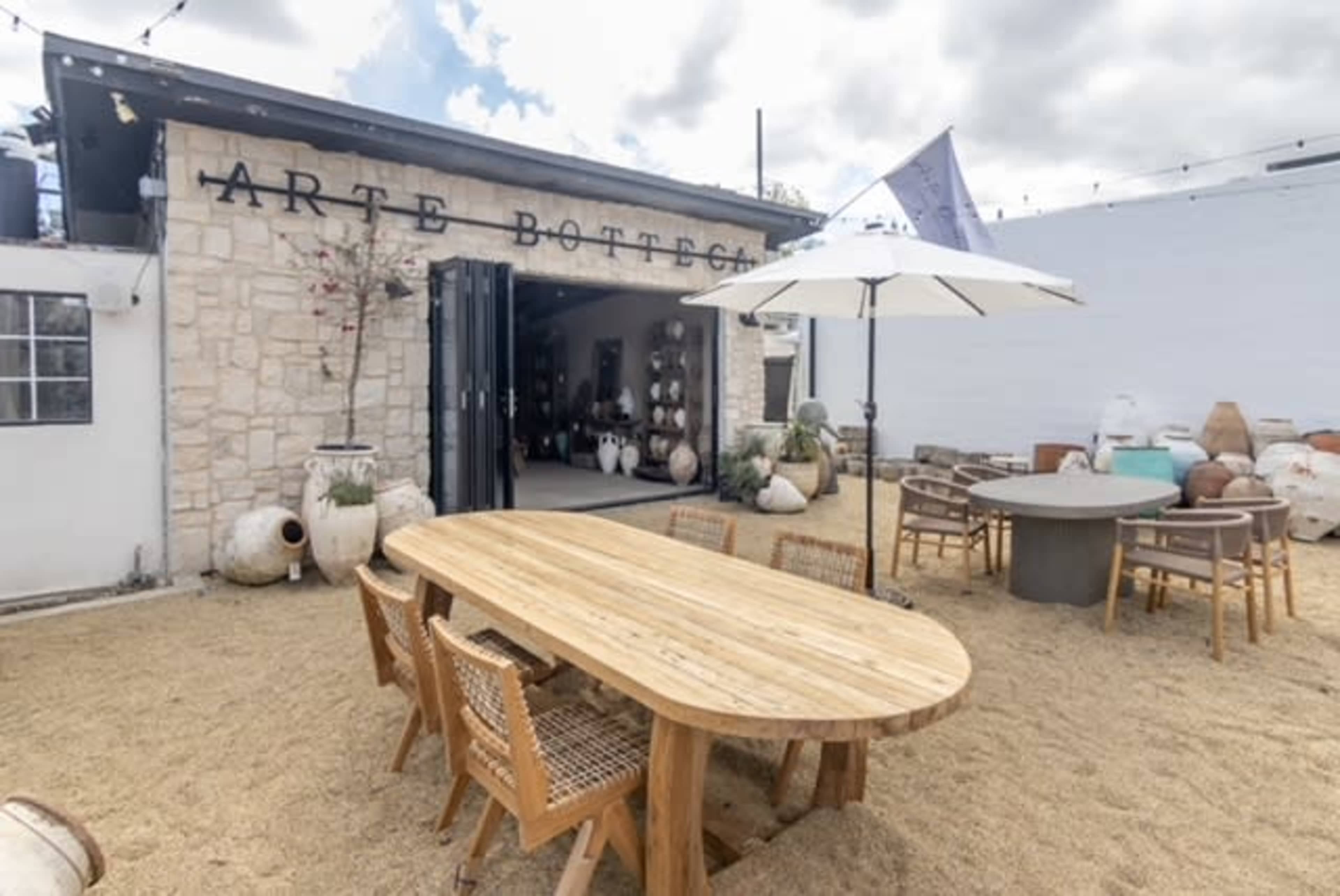 An outdoor dining area featuring a large wooden table surrounded by wicker chairs, set against a backdrop of a building with the sign "Arte Bottega" and various ceramic pots.