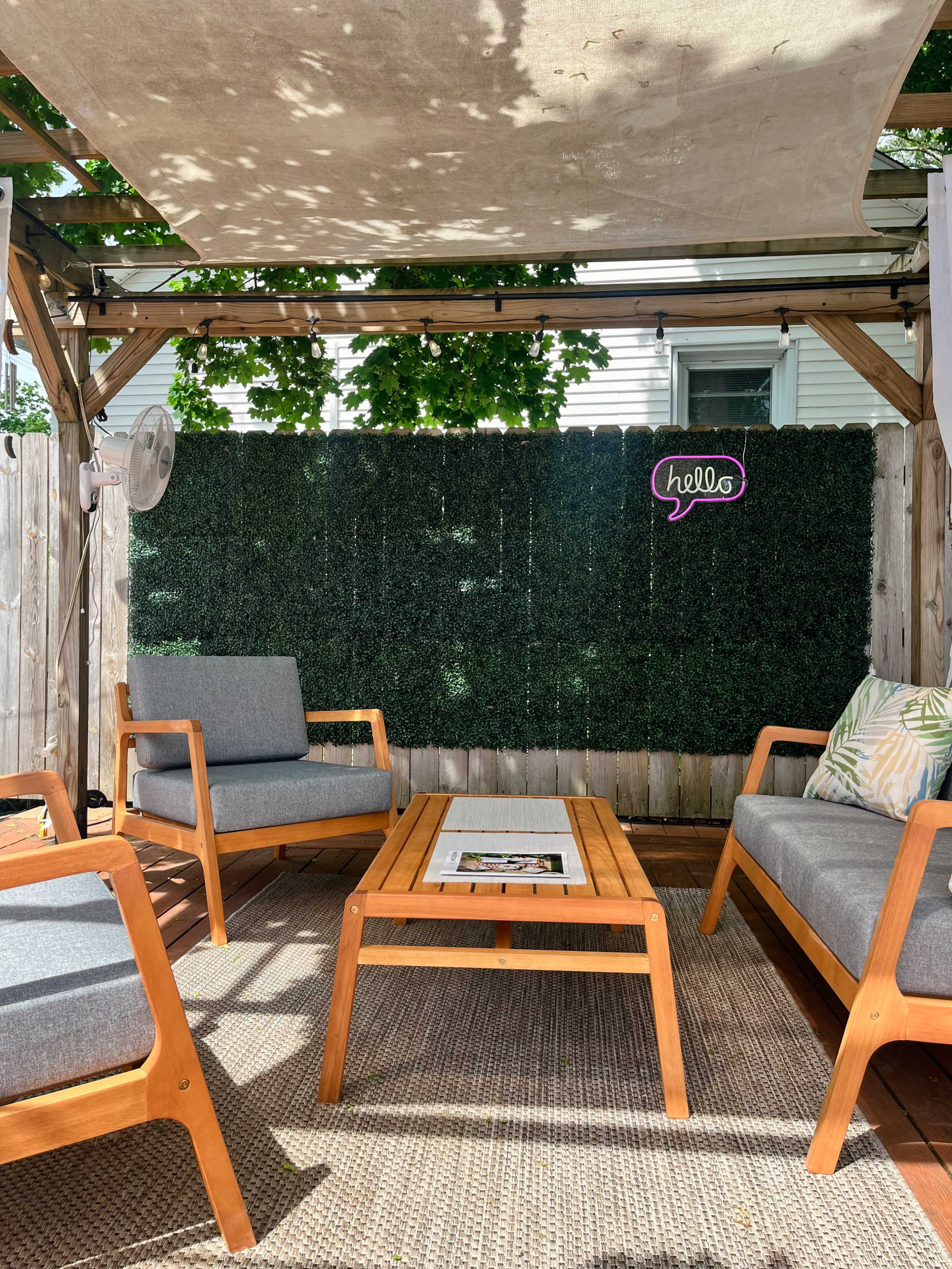 A shaded outdoor seating area with wooden furniture, including two chairs and a table, surrounded by a green wall and a decorative "hello" sign.