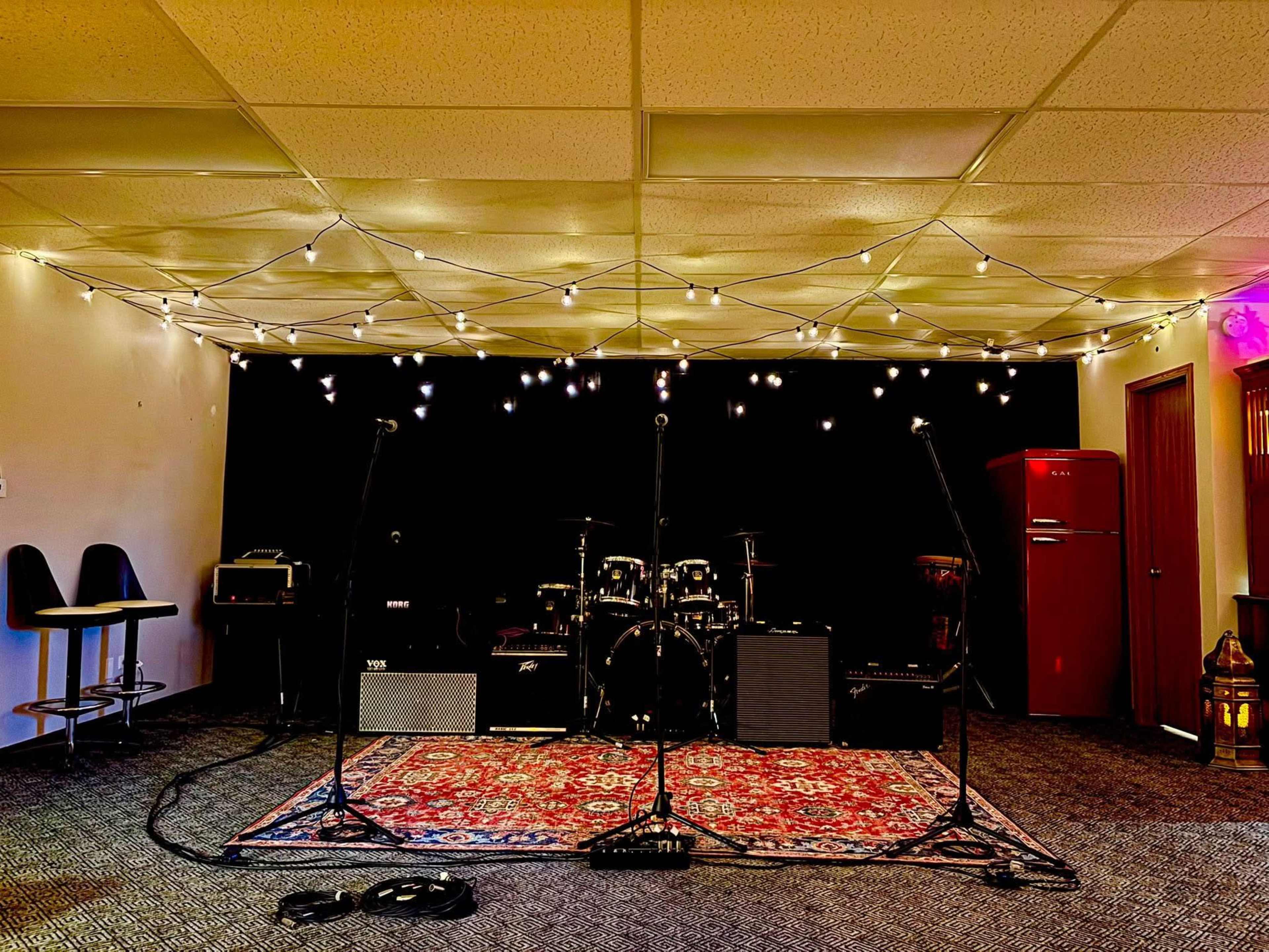 The image shows a music rehearsal space with microphones set up on stands, amplifiers positioned on the floor, and a drum kit in the background, all beneath a canopy of string lights.