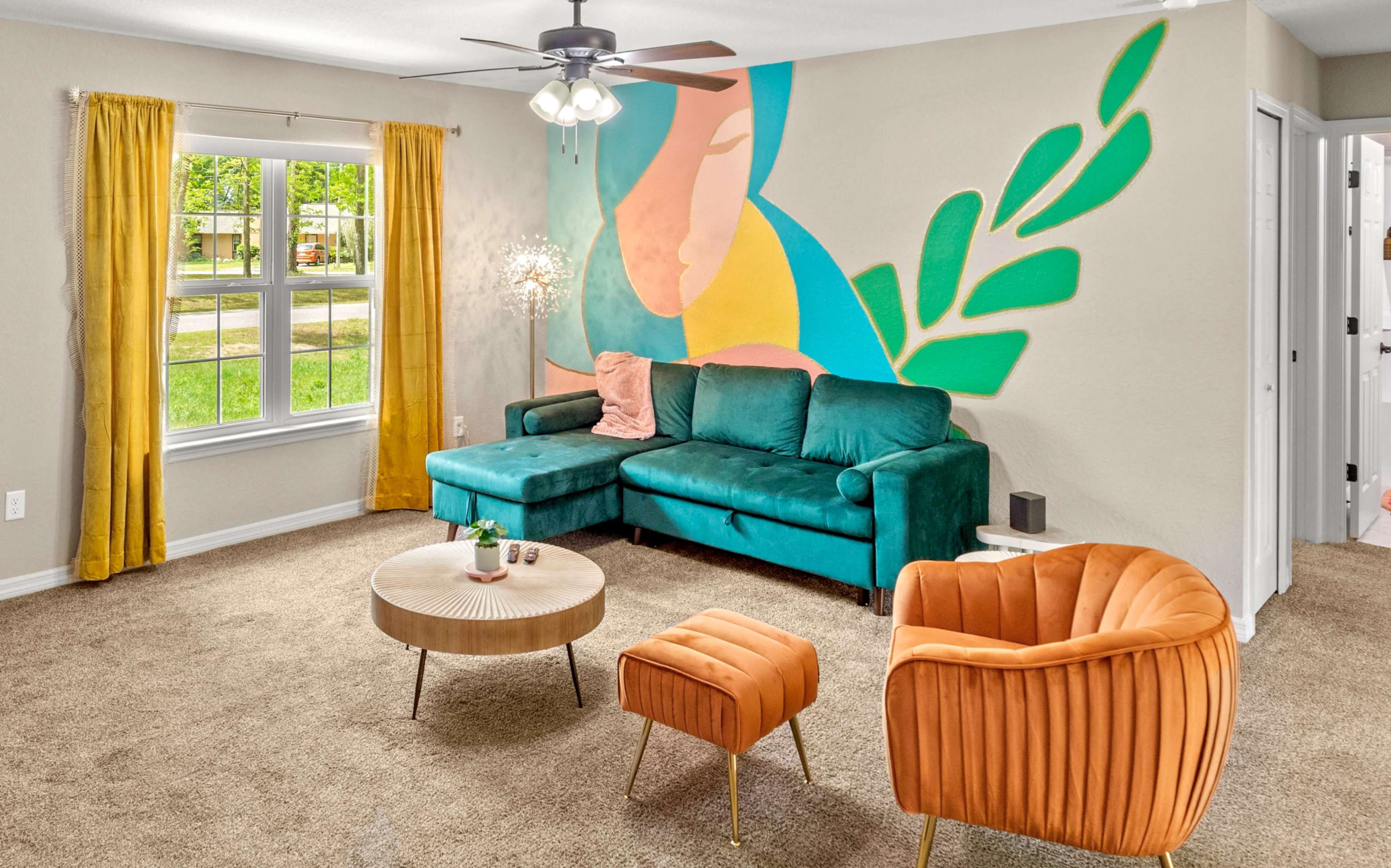 A living room with a green couch, an orange chair, a circular coffee table, and a colorful mural on the wall.
