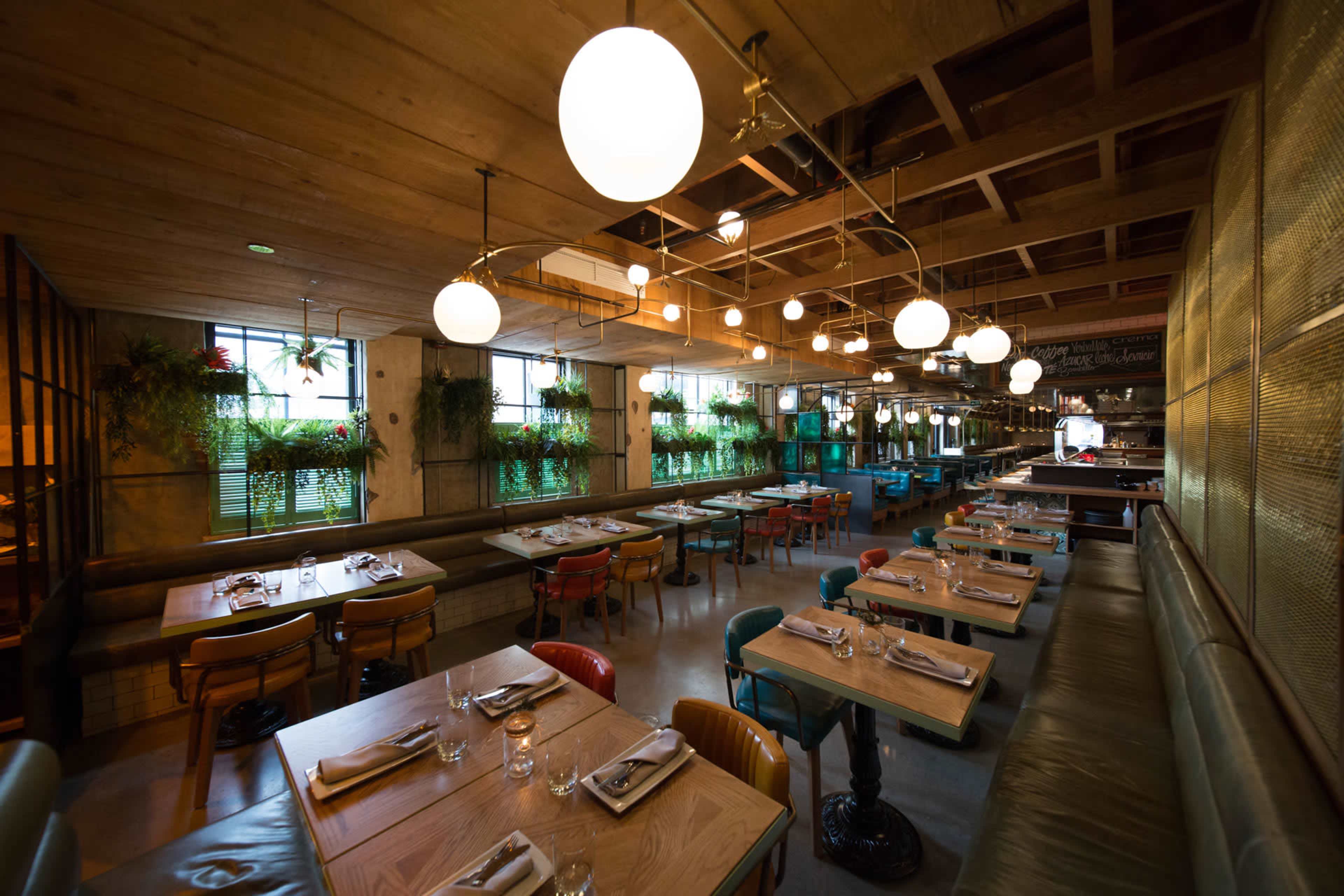 The image shows a modern restaurant interior with wooden tables, colorful chairs, and pendant lighting, arranged in a spacious layout.