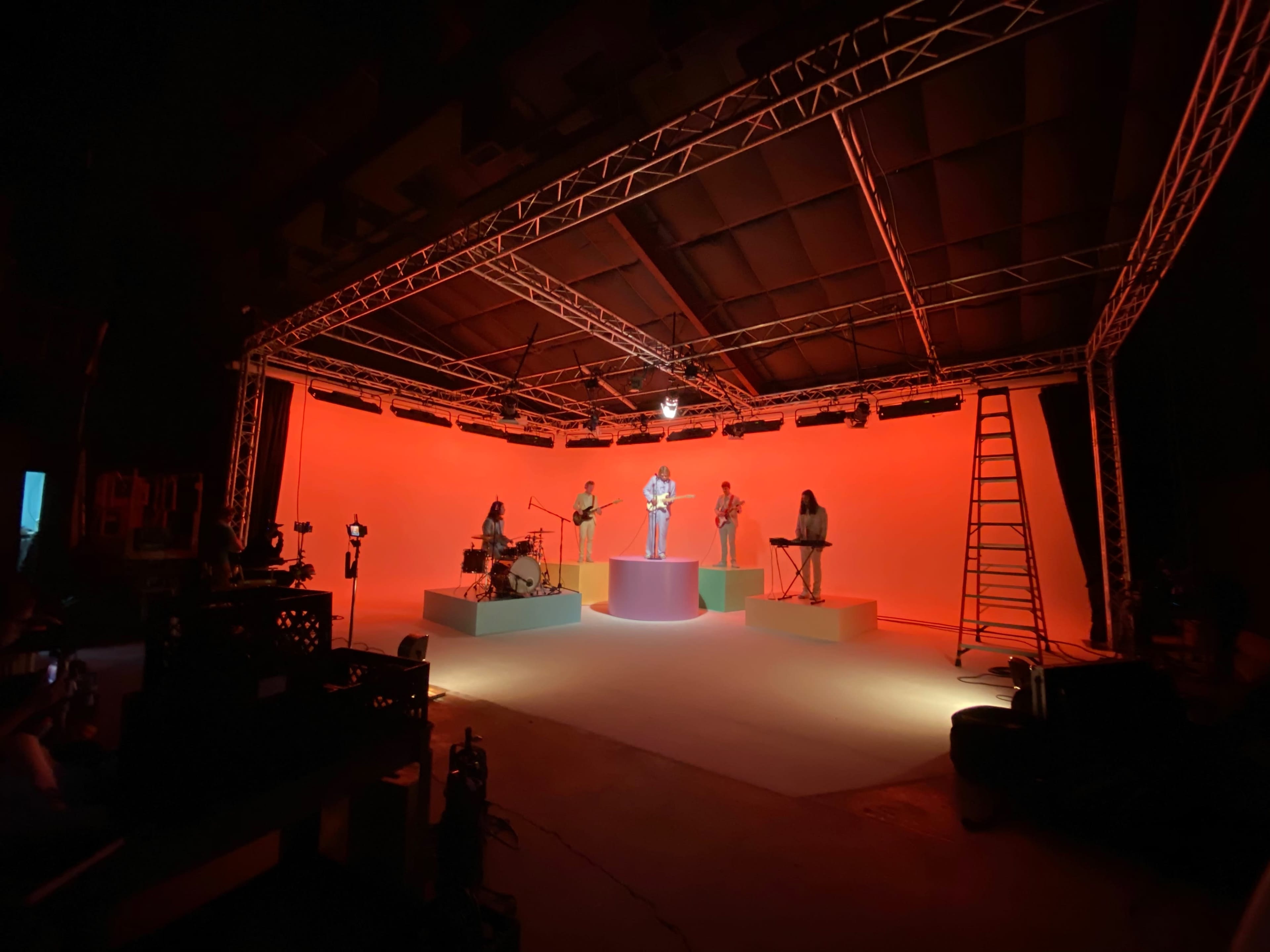 A musical performance is taking place on a brightly lit stage with several musicians standing on colorful platforms against an orange background.