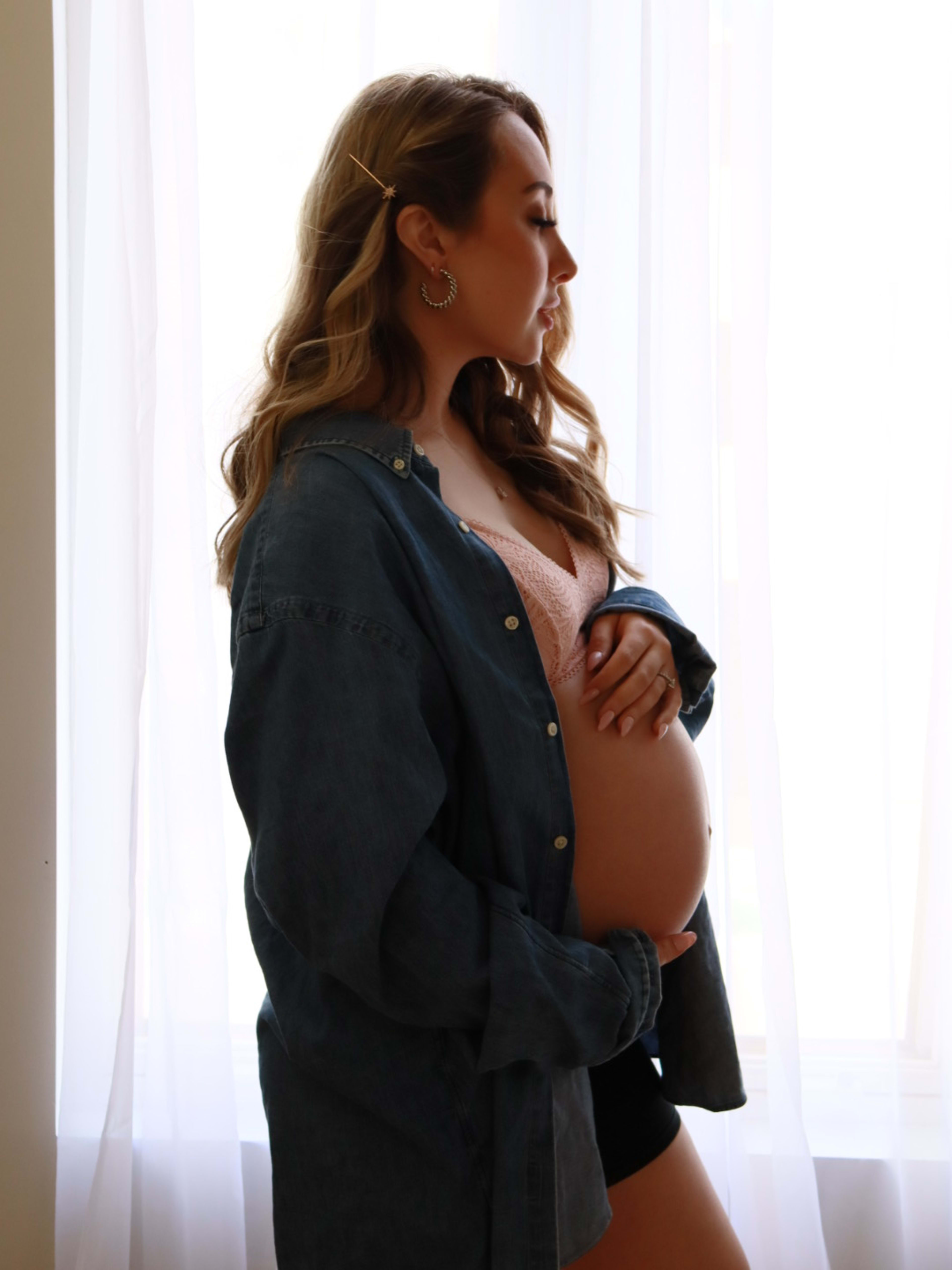 A pregnant woman peacefully standing by the window.