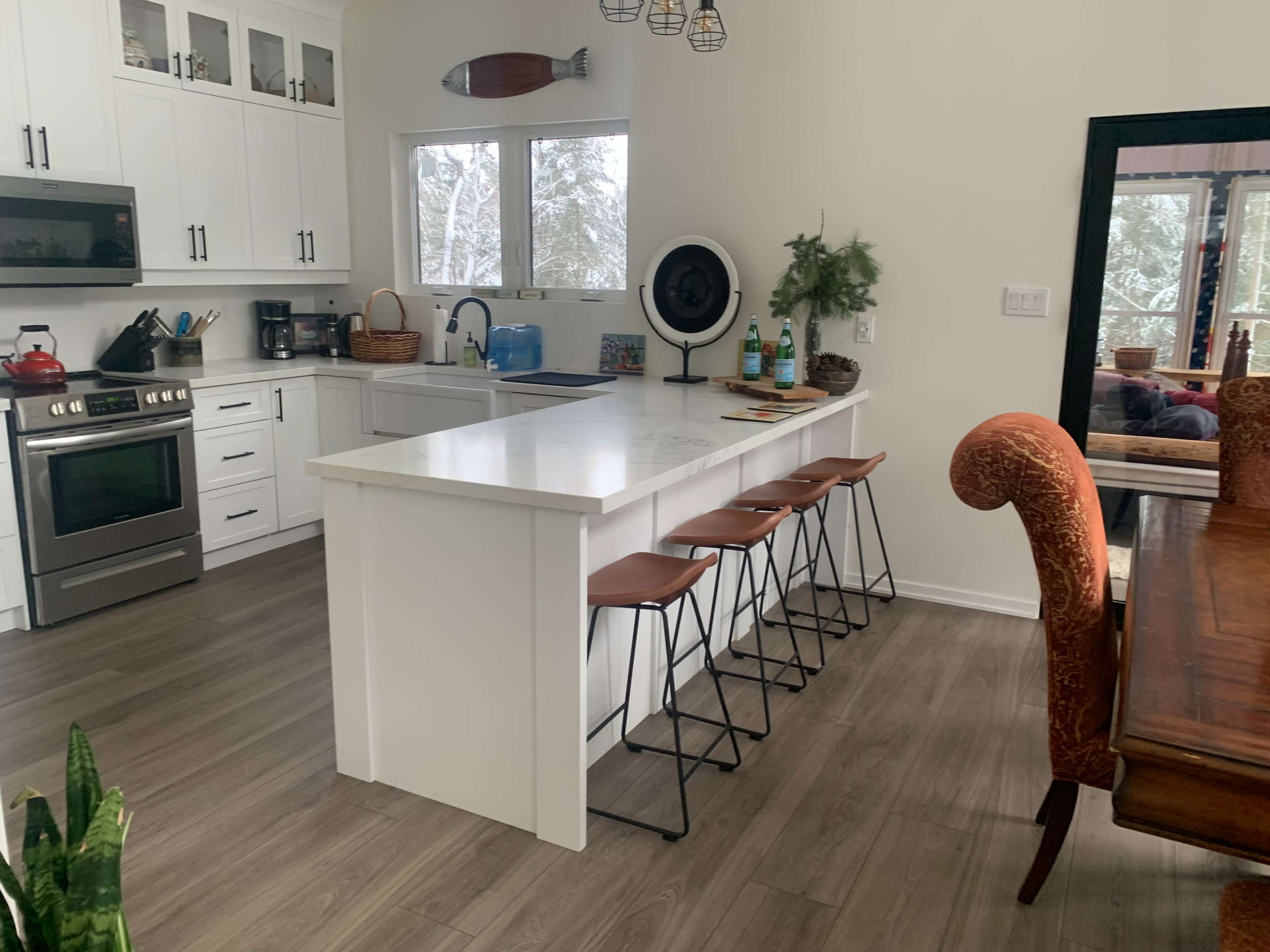 A modern kitchen features a central island with four high stools, stainless steel appliances, and large windows allowing natural light in.