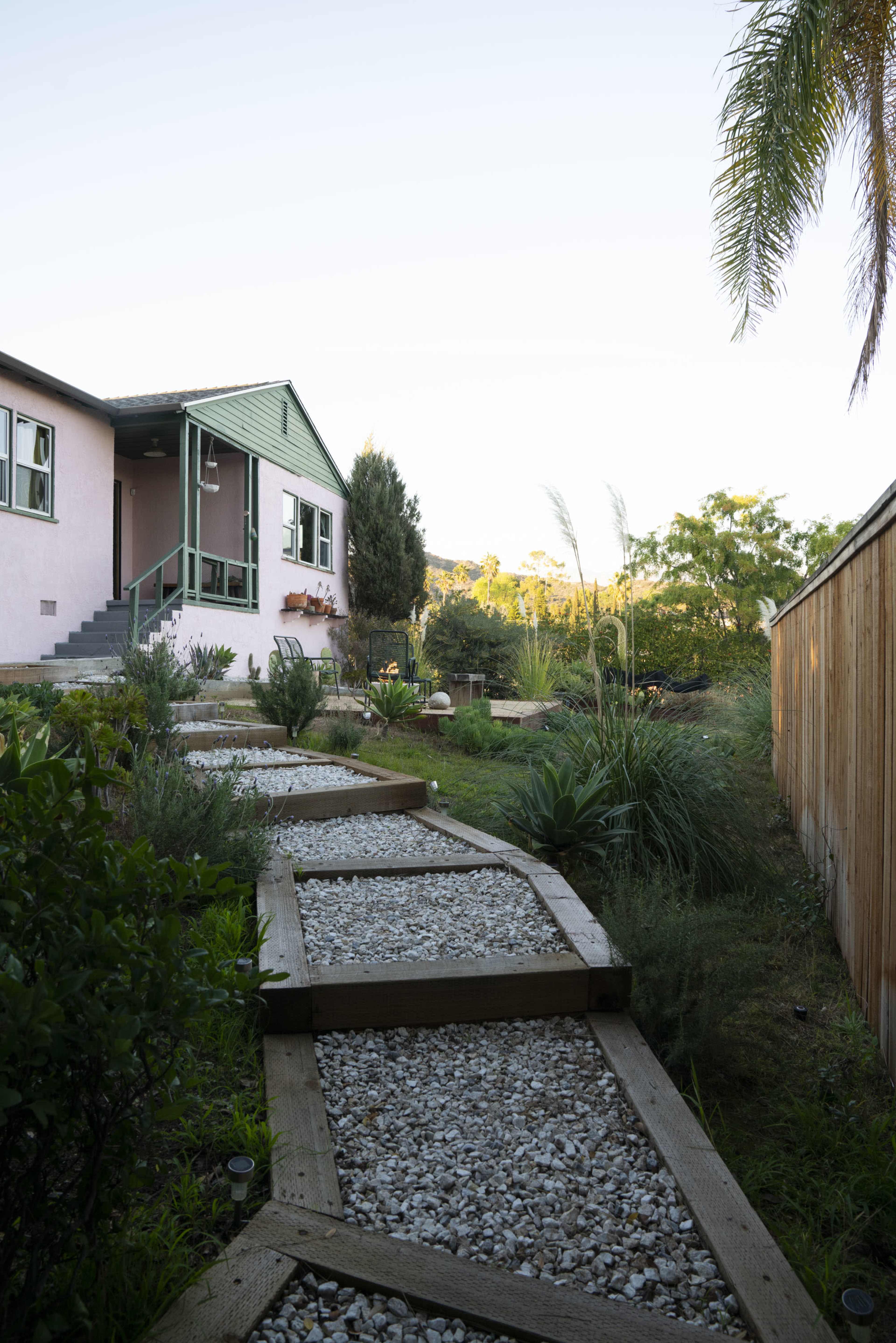 A wooden walkway lined with gravel leads to a house surrounded by various plants and greenery.
