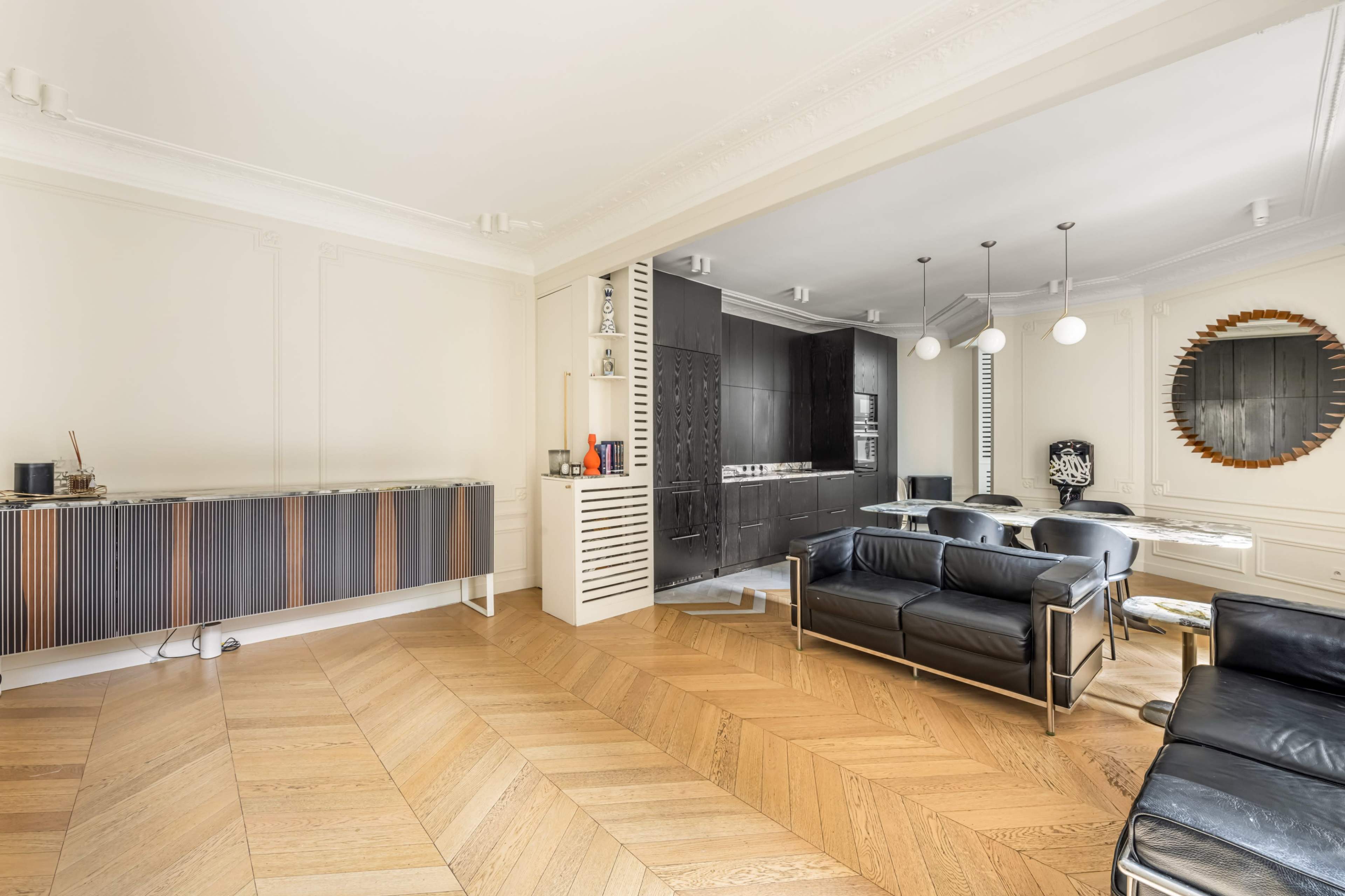 A modern living room features a wooden herringbone floor, black leather seating, and a sleek kitchen area with dark cabinetry.