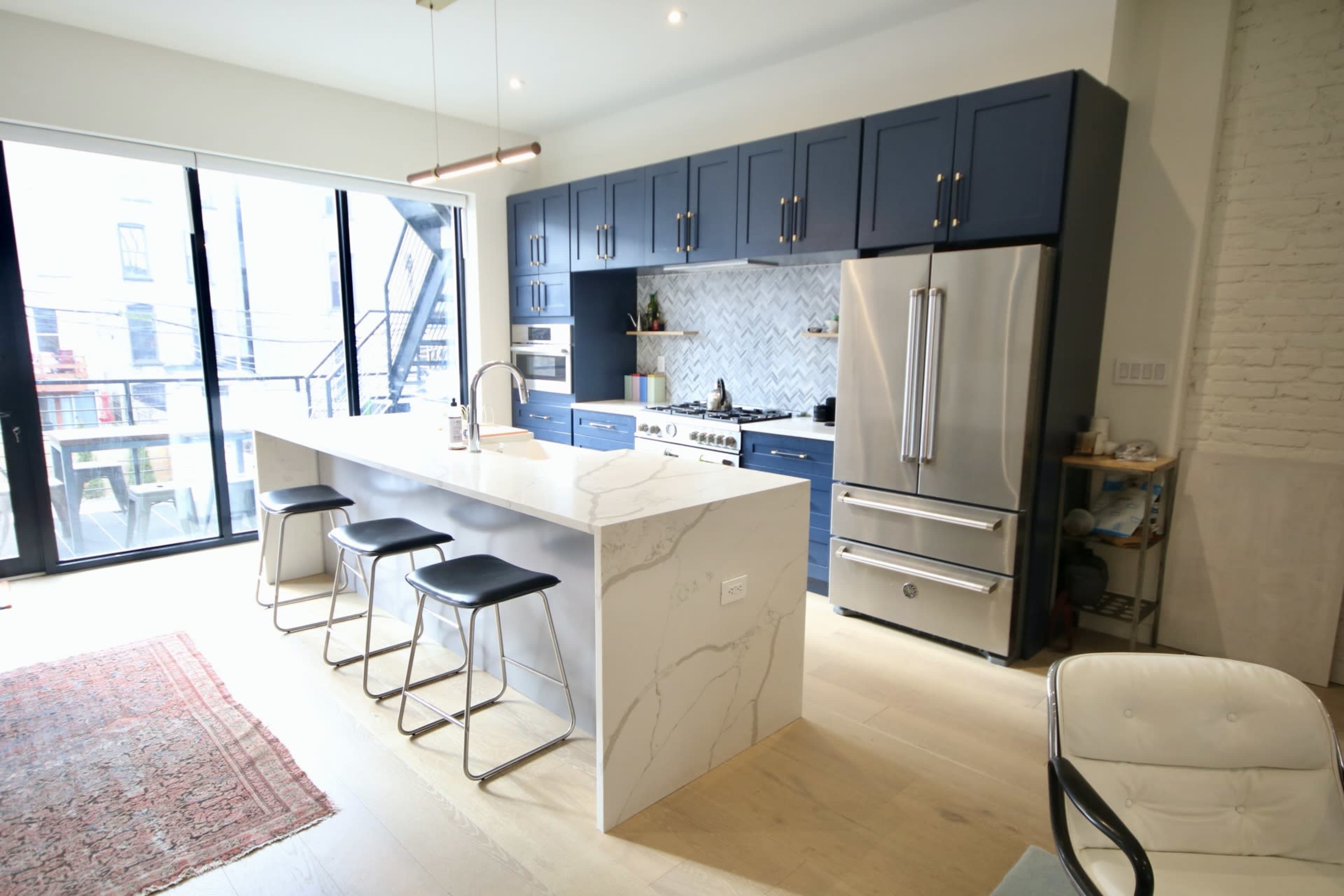 A modern kitchen features a large island with a white marble countertop, navy cabinets, stainless steel appliances, and a spacious layout with large windows.