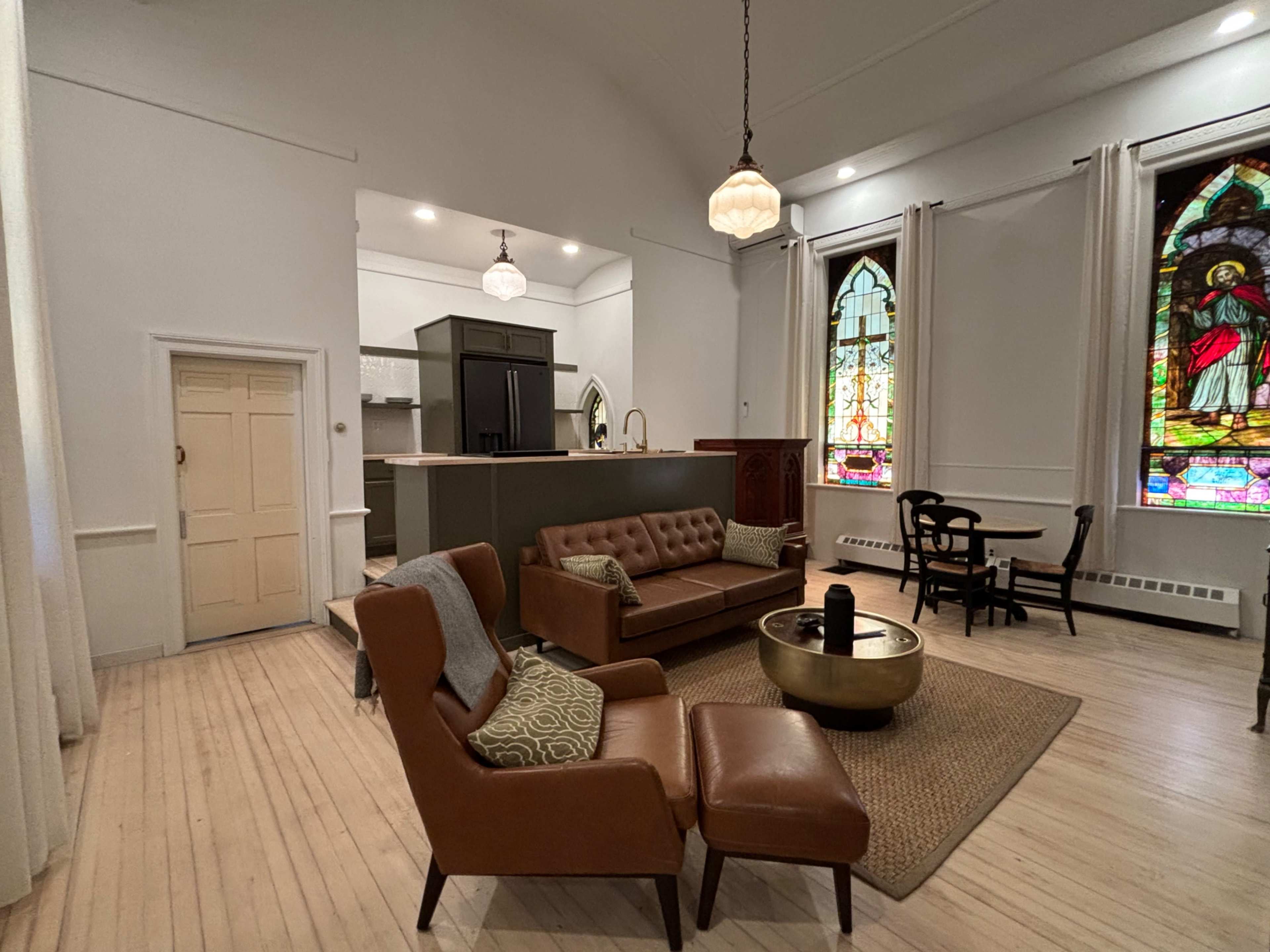 A renovated living space featuring a brown leather sofa, an armchair, a round coffee table, and a stained glass window, all set in a bright room with wooden floors and a modern kitchen area visible in the background.