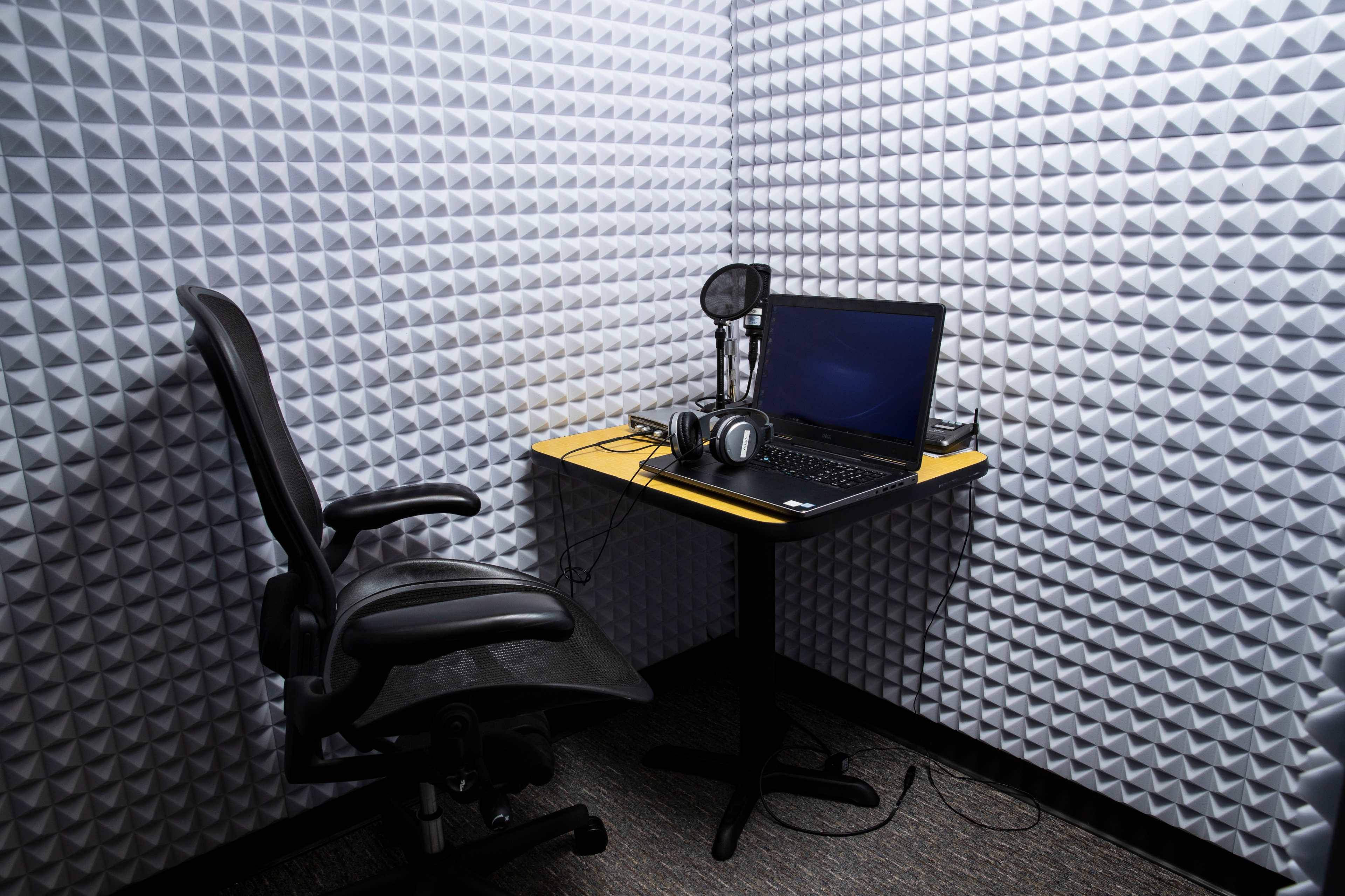 A small recording studio features a desk with a laptop, headphones, and a microphone, surrounded by soundproof foam panels.
