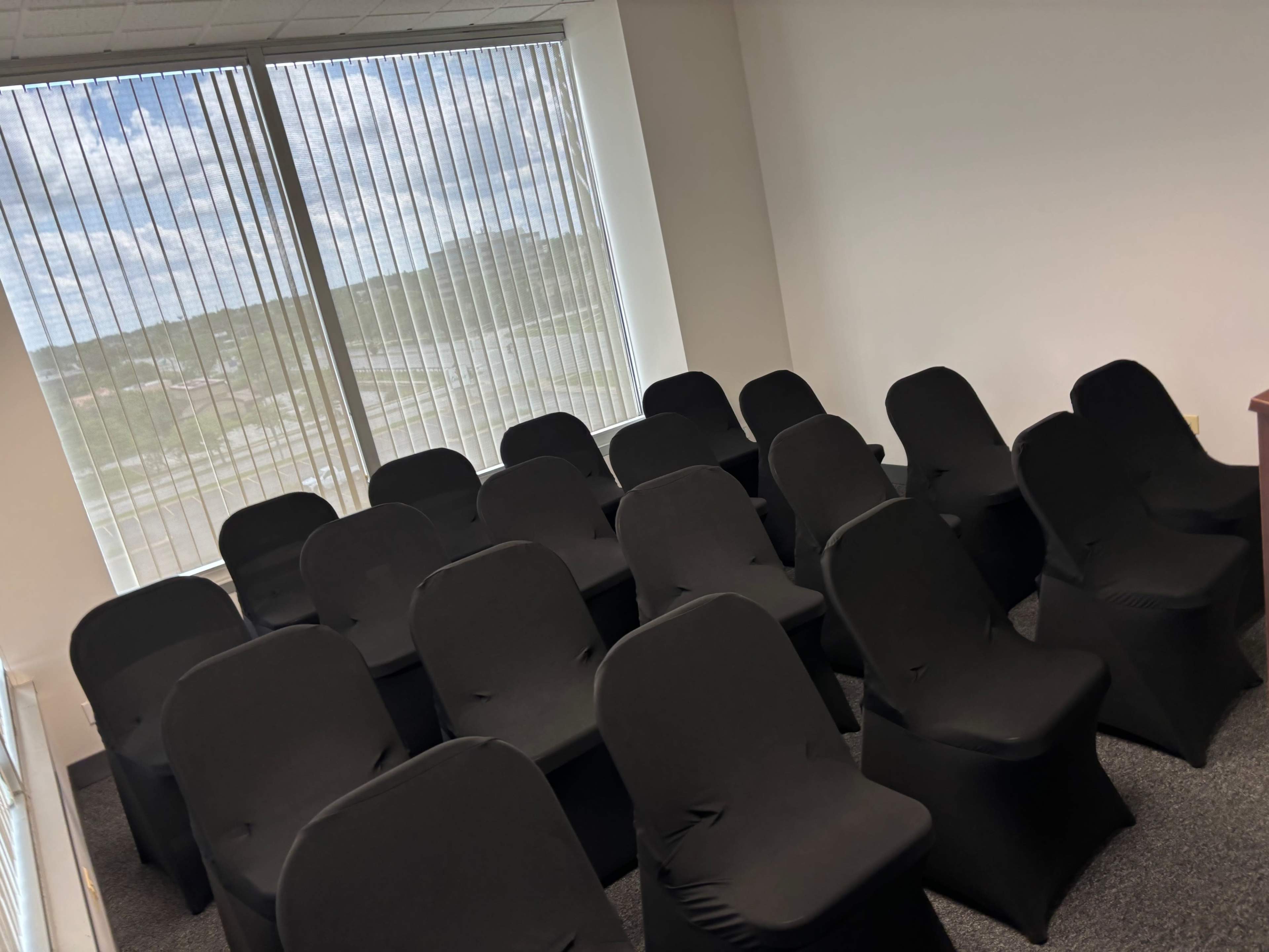 A row of covered black chairs is arranged in a meeting room with large windows allowing natural light to enter.