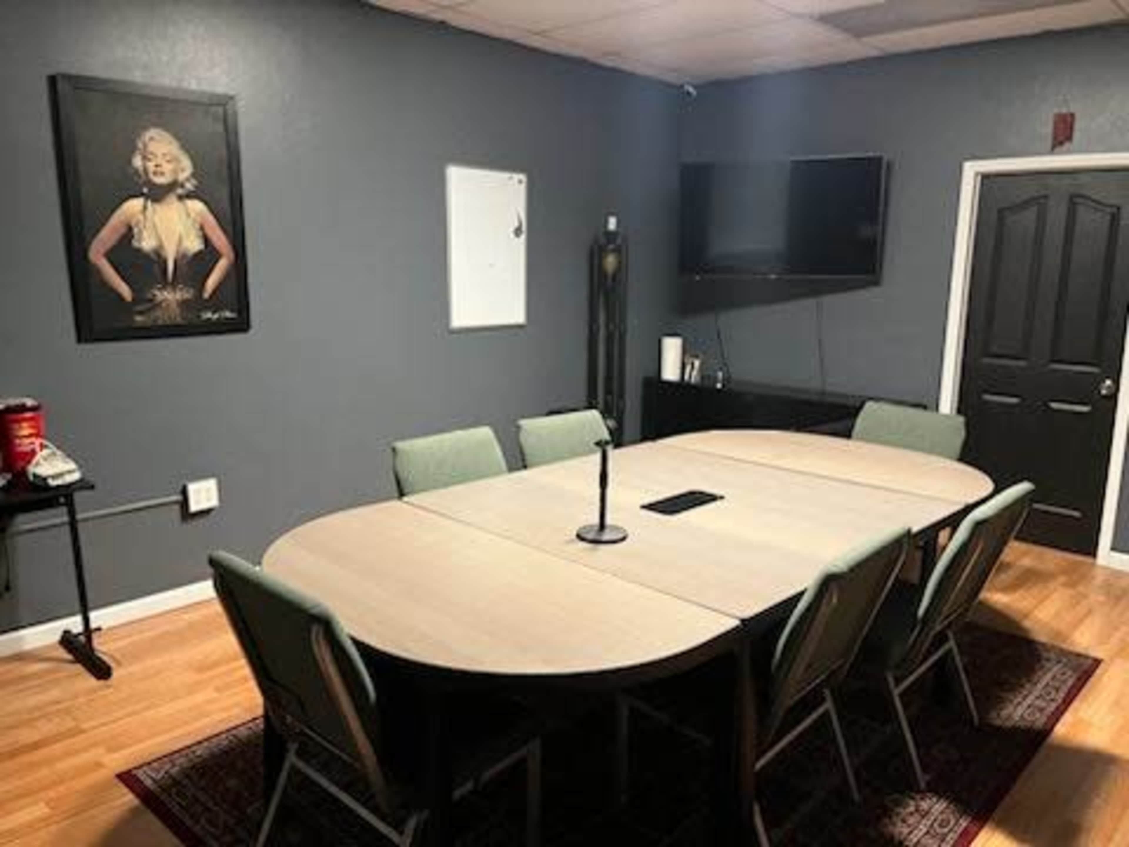 A conference room with a large oval table surrounded by eight green chairs, a wall-mounted TV, and a framed poster on the wall.
