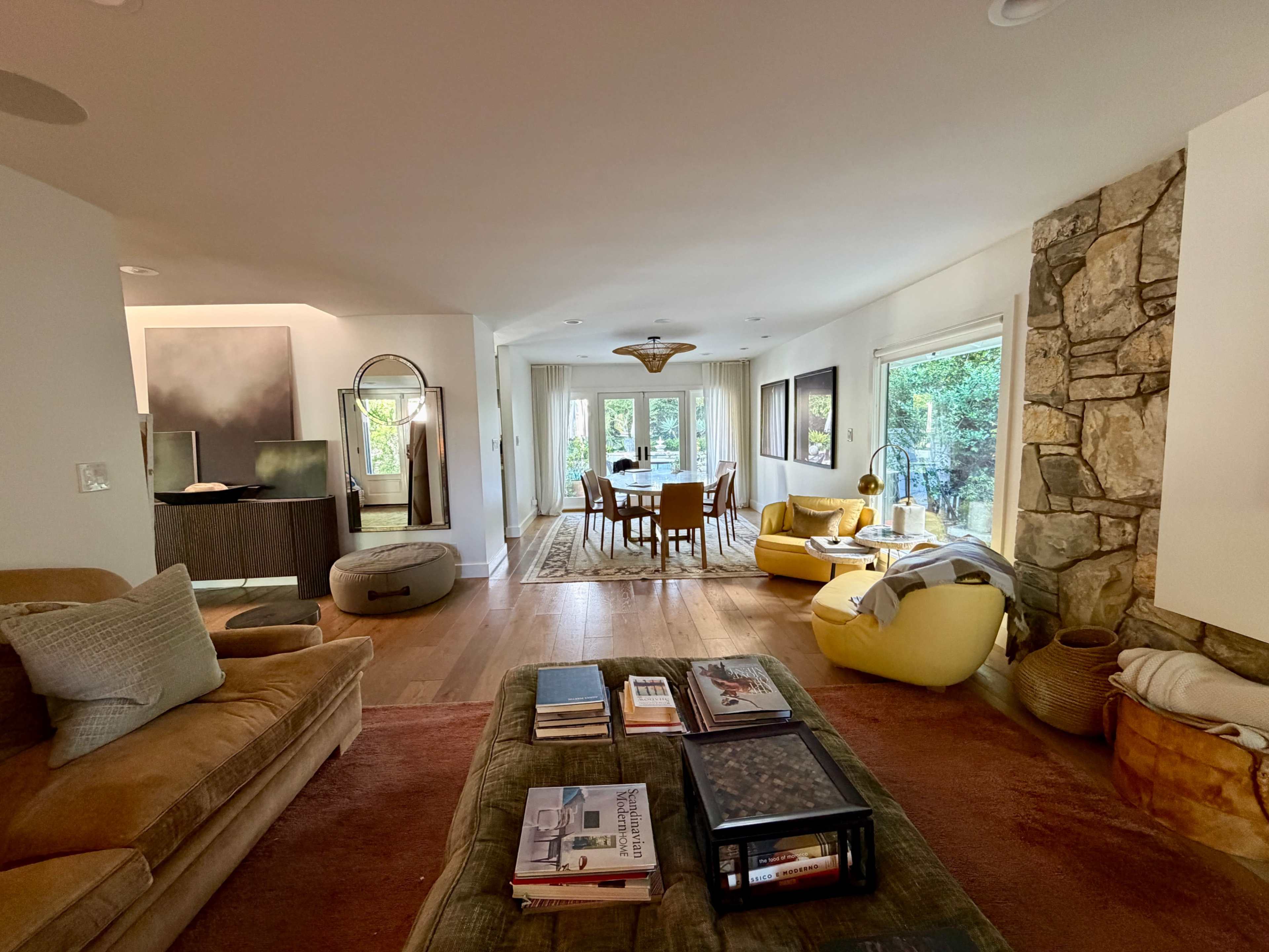 The image shows a cozy living room with a mix of comfortable seating, a wooden dining table in the background, and large windows allowing natural light to enter.