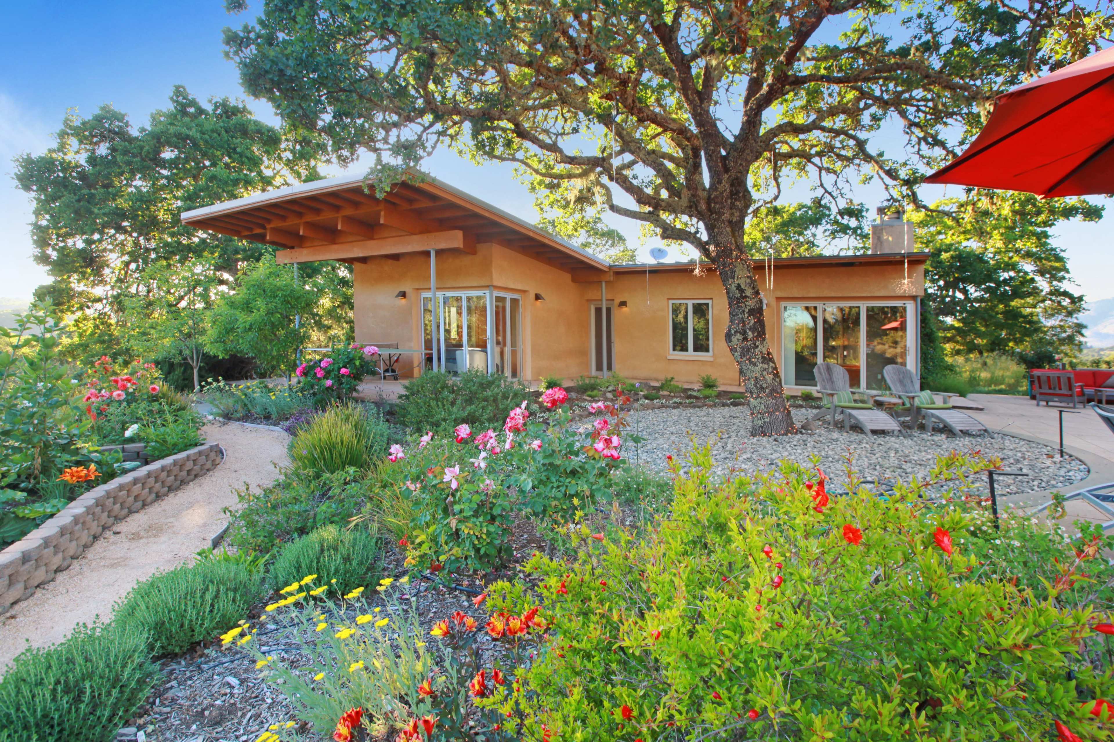 A modern, single-story home surrounded by landscaped gardens filled with various flowers and plants under a large tree.