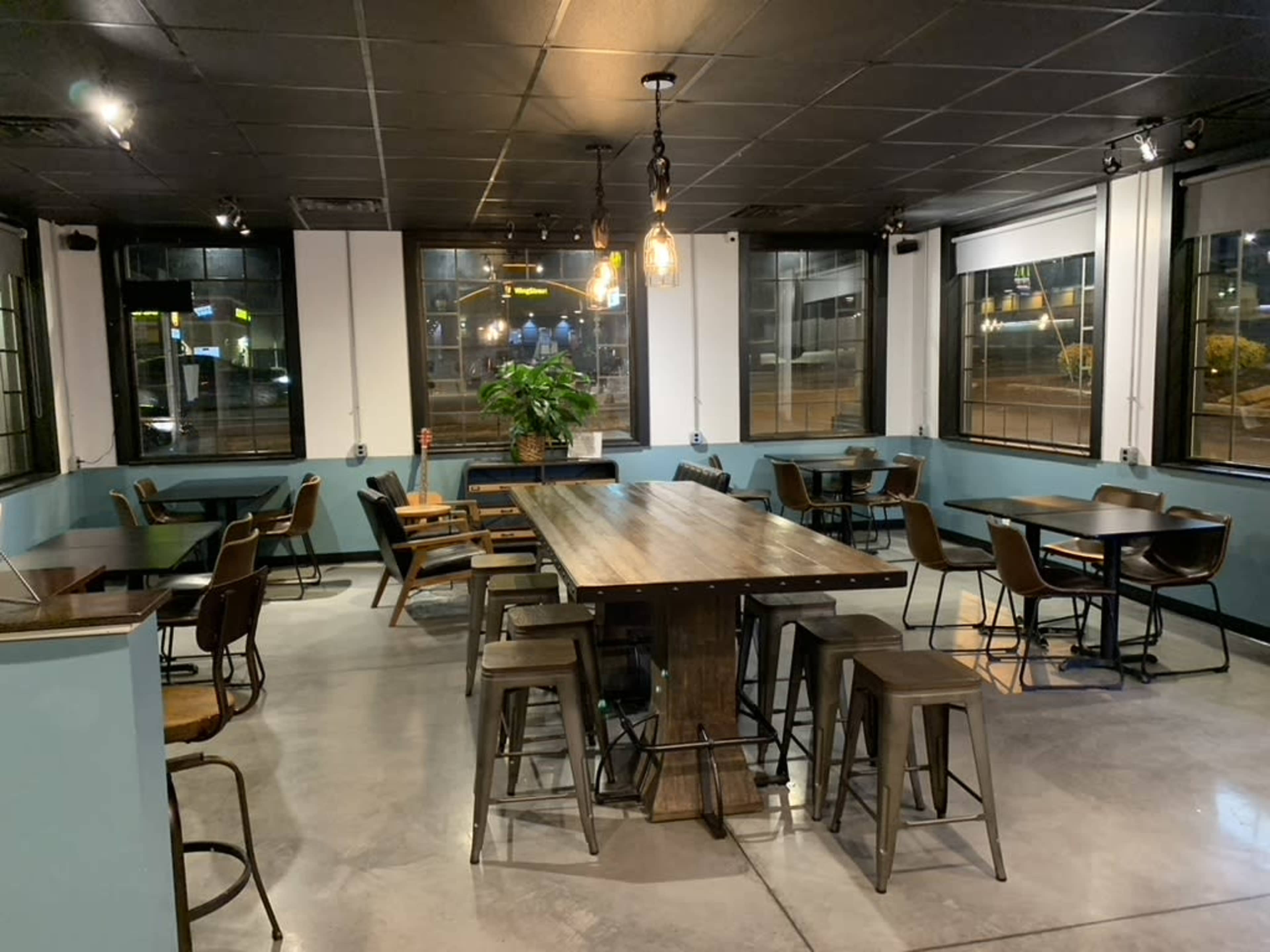 The image shows a modern café interior with a long communal table, several smaller tables, and large windows facing the street.