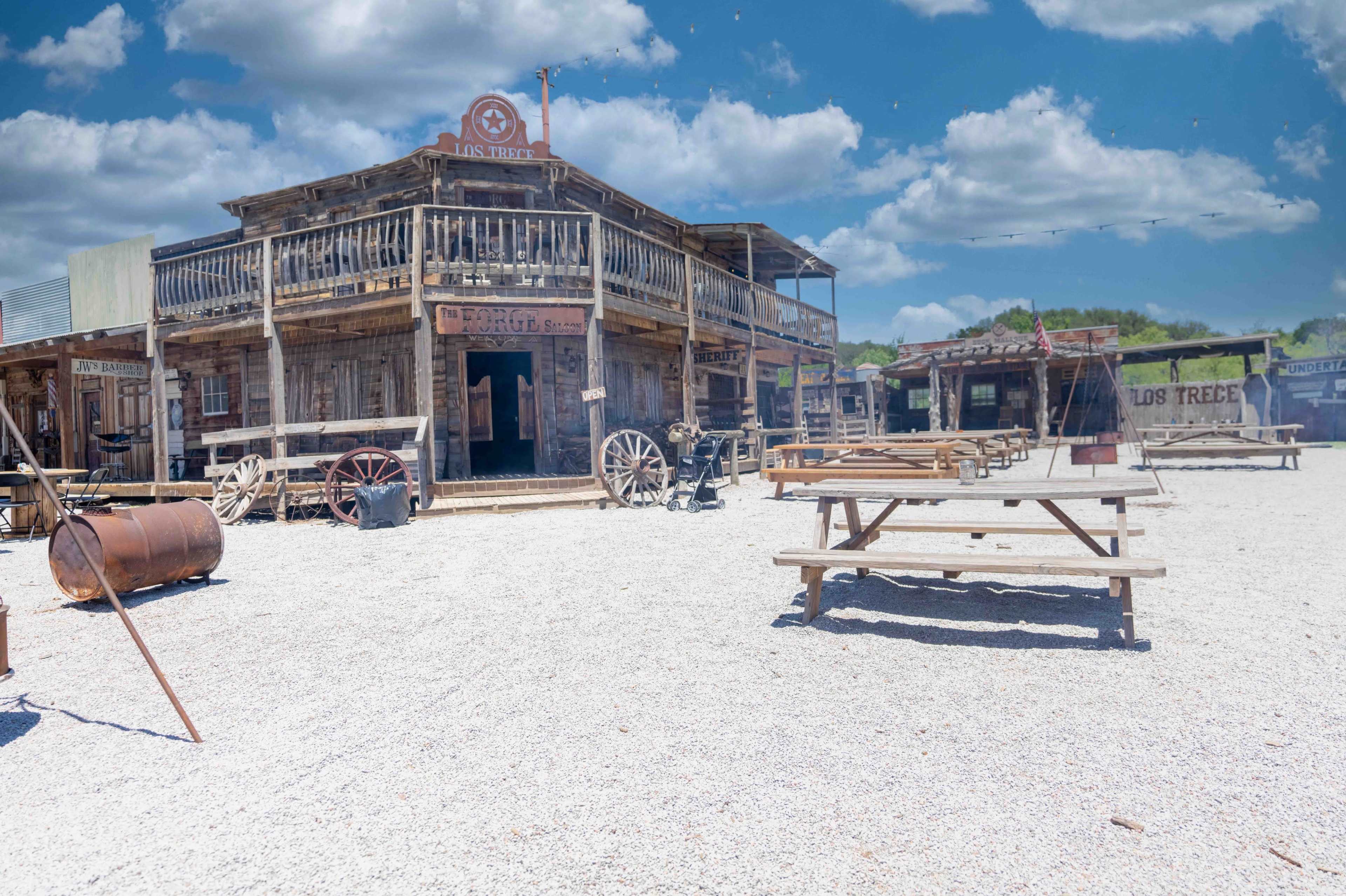 The image depicts a western-style town with wooden buildings, wagon wheels, and picnic tables set on a gravel surface under a partly cloudy sky.
