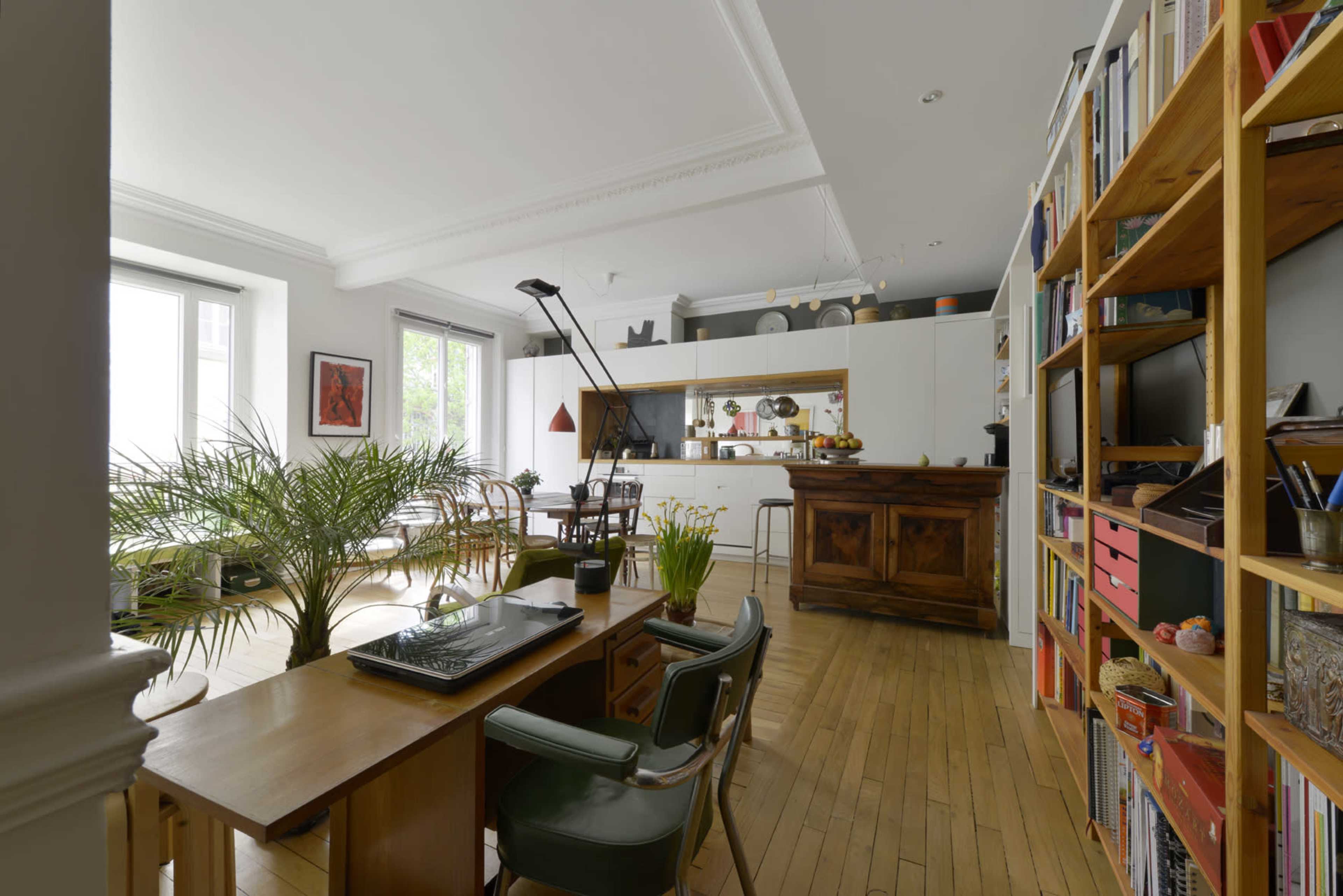 A modern living space features a wooden dining table, a kitchen area with white cabinetry, and a wooden bookshelf filled with books, plants, and decorative items.