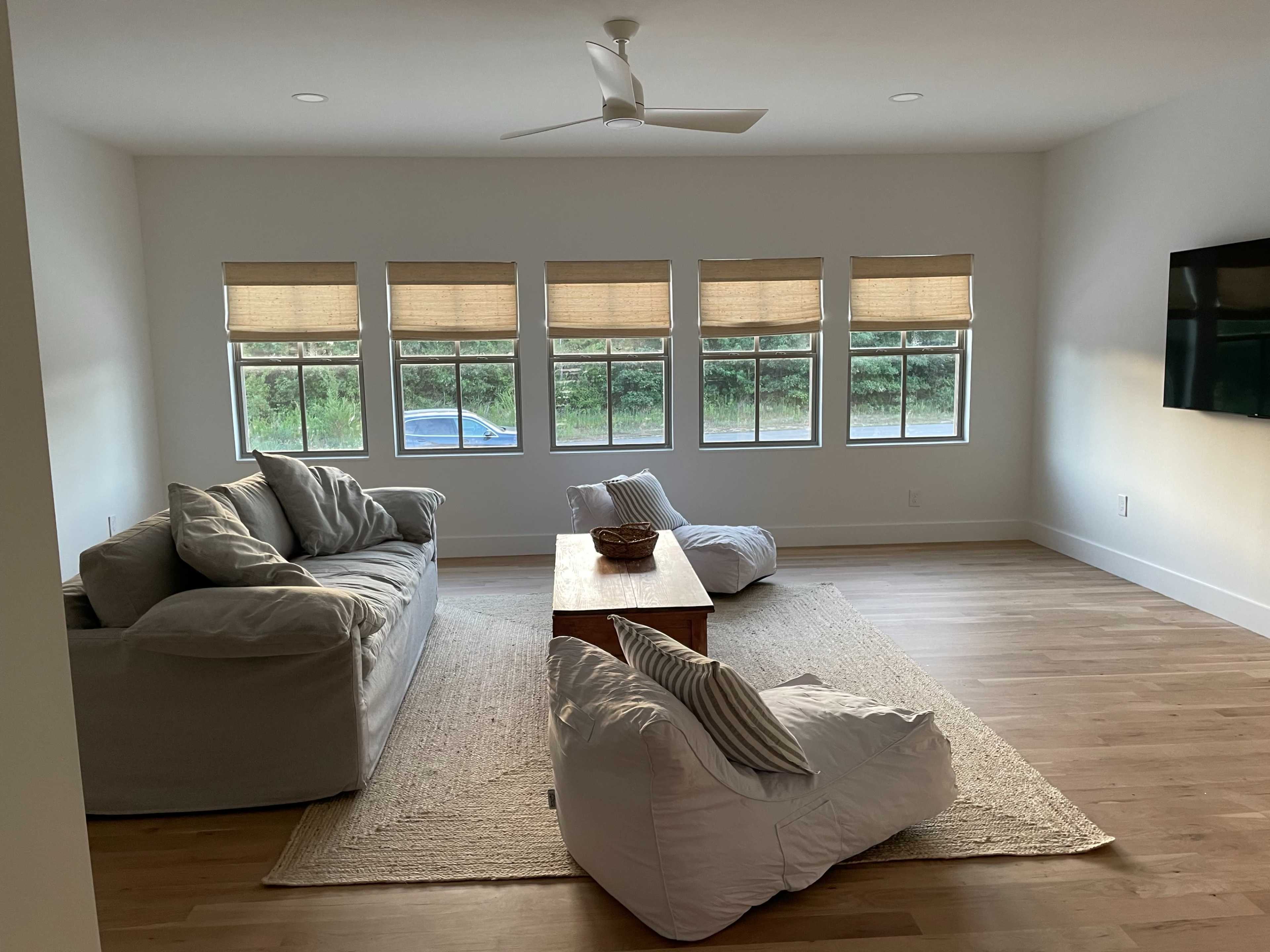 A spacious living room features two sofas, a wooden coffee table, and bean bag chairs, with large windows allowing natural light to illuminate the space.