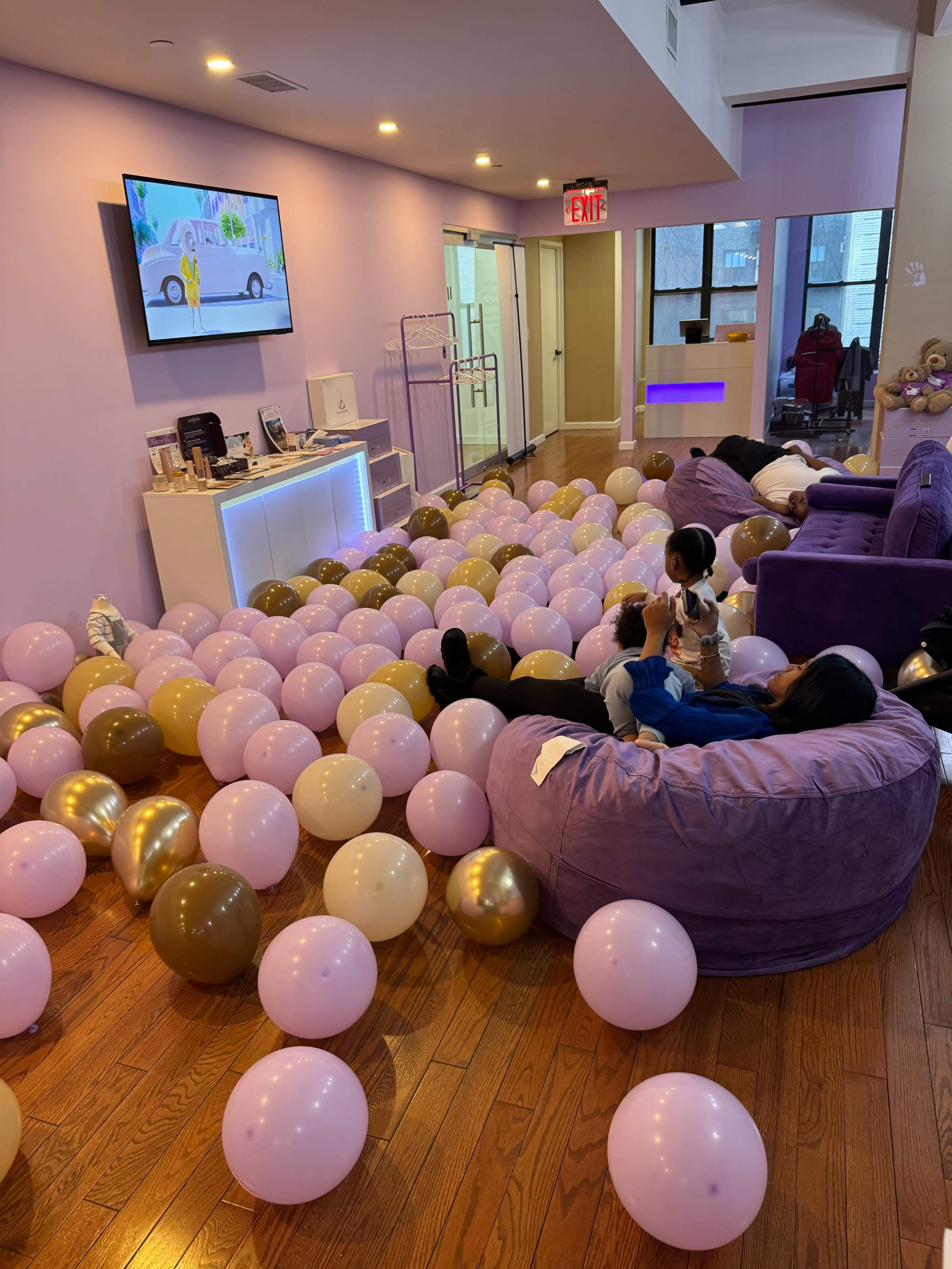 A large room filled with numerous pastel-colored balloons covers the floor, while a television displays a cartoon on the wall.