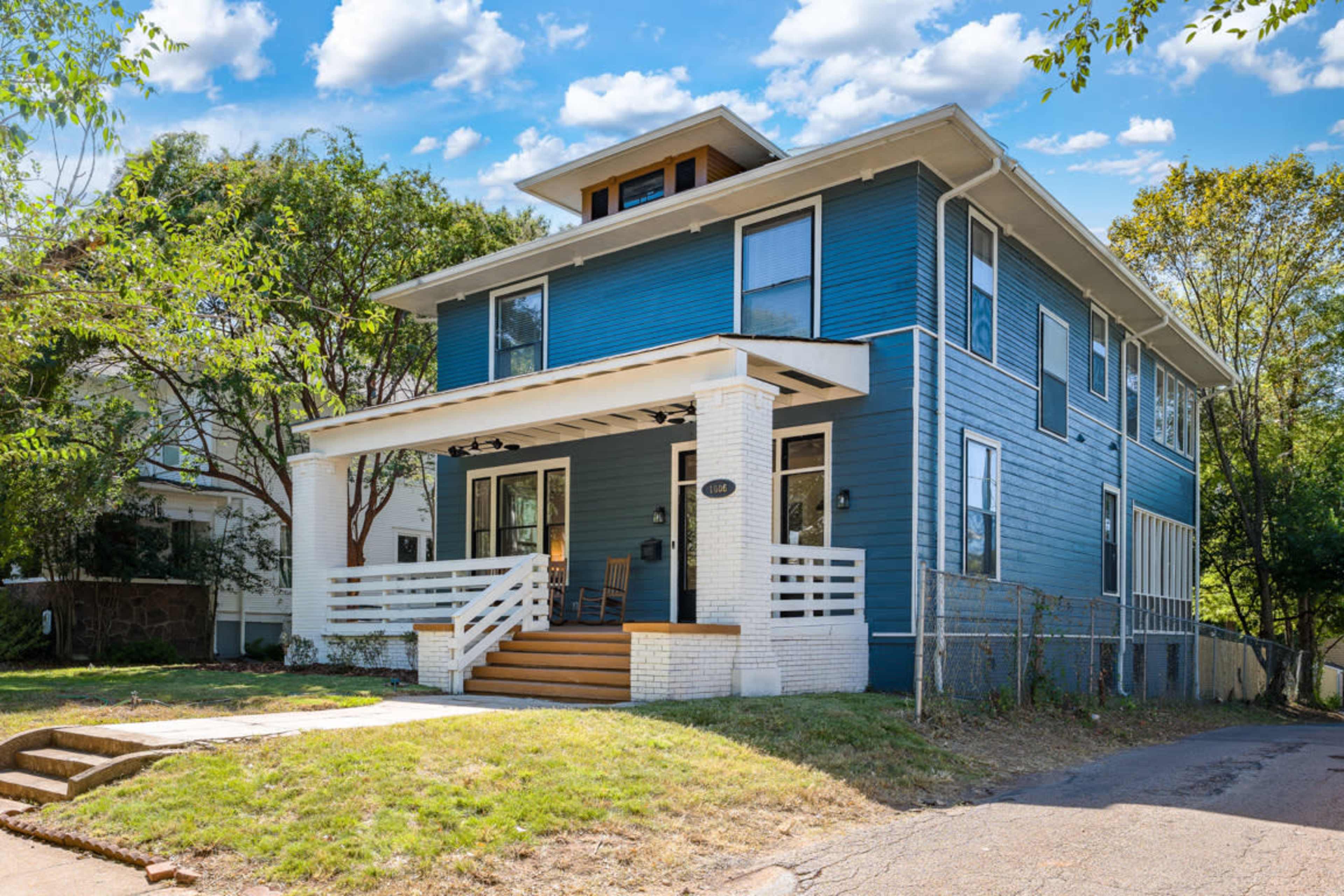 A three-story blue house with a front porch, white trim, and a tree-filled lawn on a quiet street.