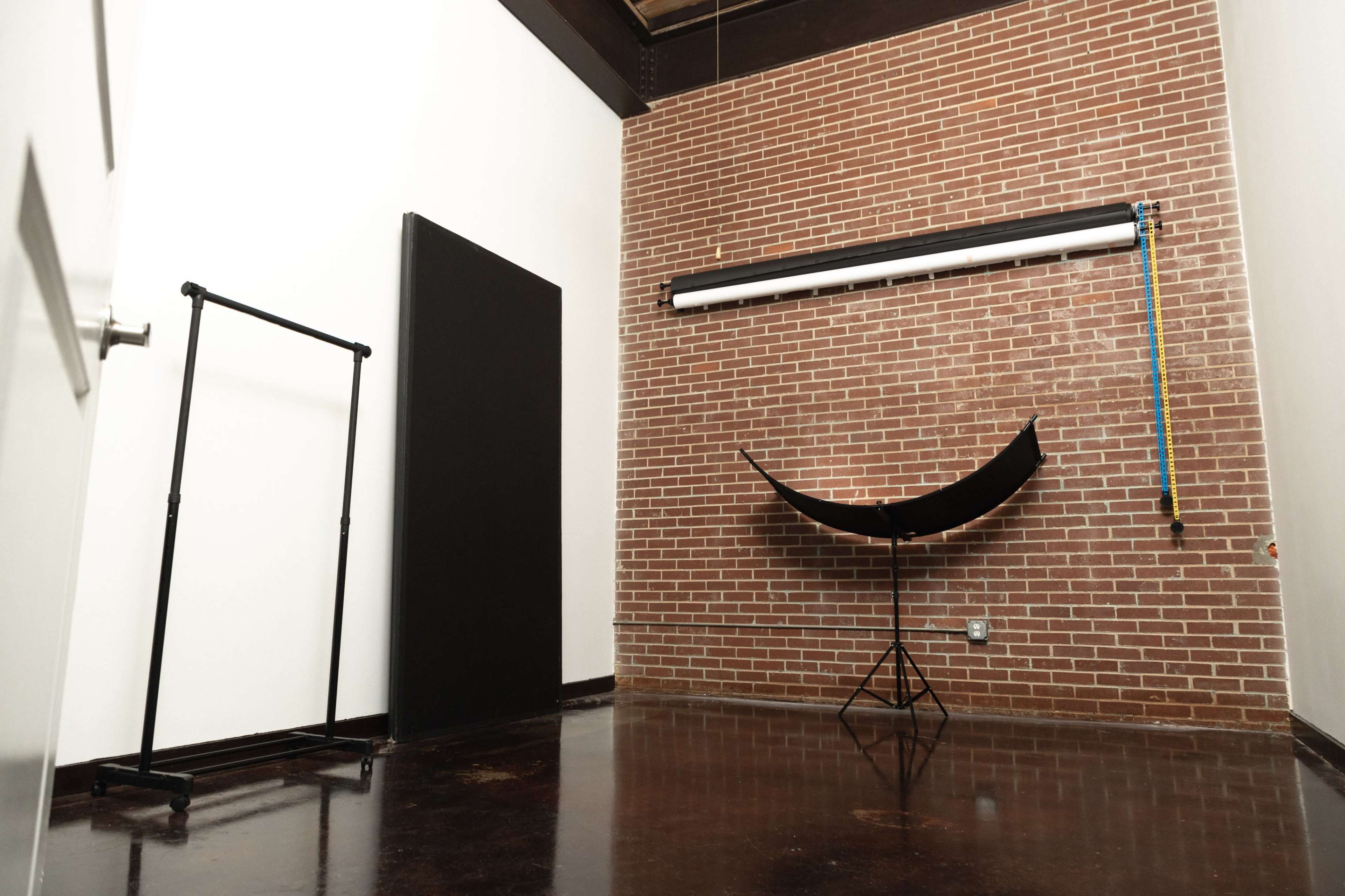 A photography studio with a brick wall, a black backdrop on a frame, a curved reflector stand, and a measuring tape hanging from a rail.