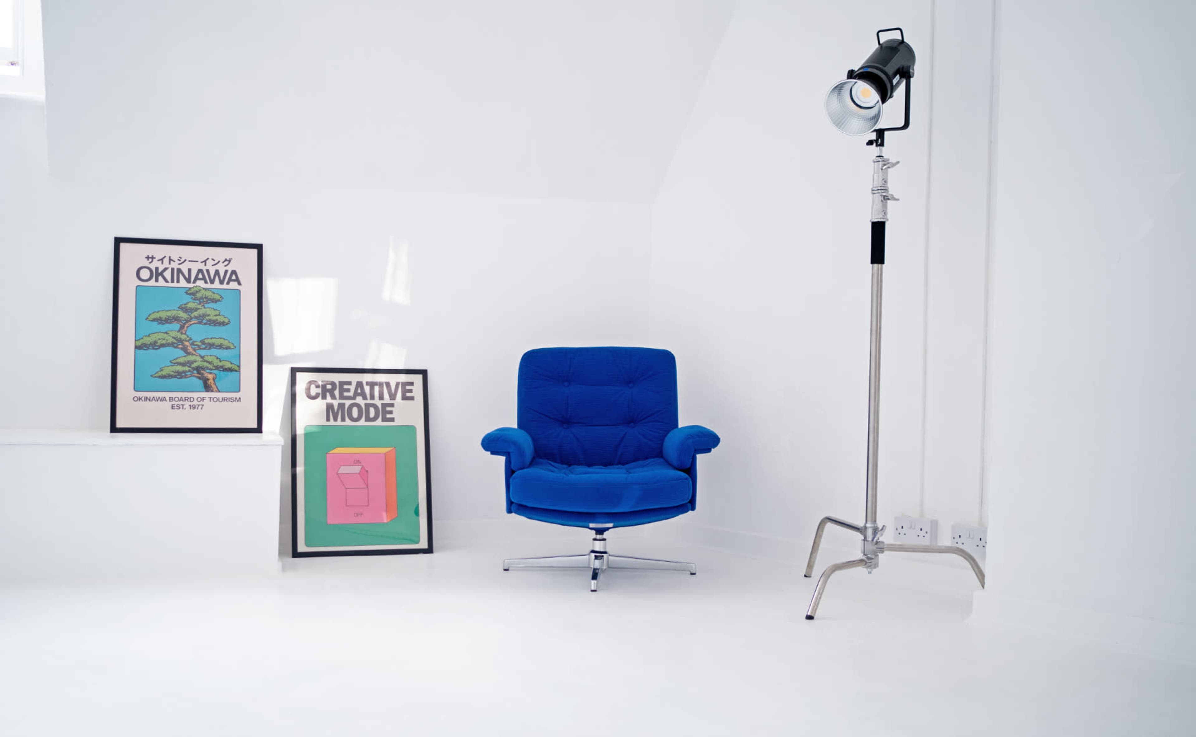 The image shows a bright, minimalist room featuring a blue armchair, framed artworks on the wall, and a studio light on a stand.