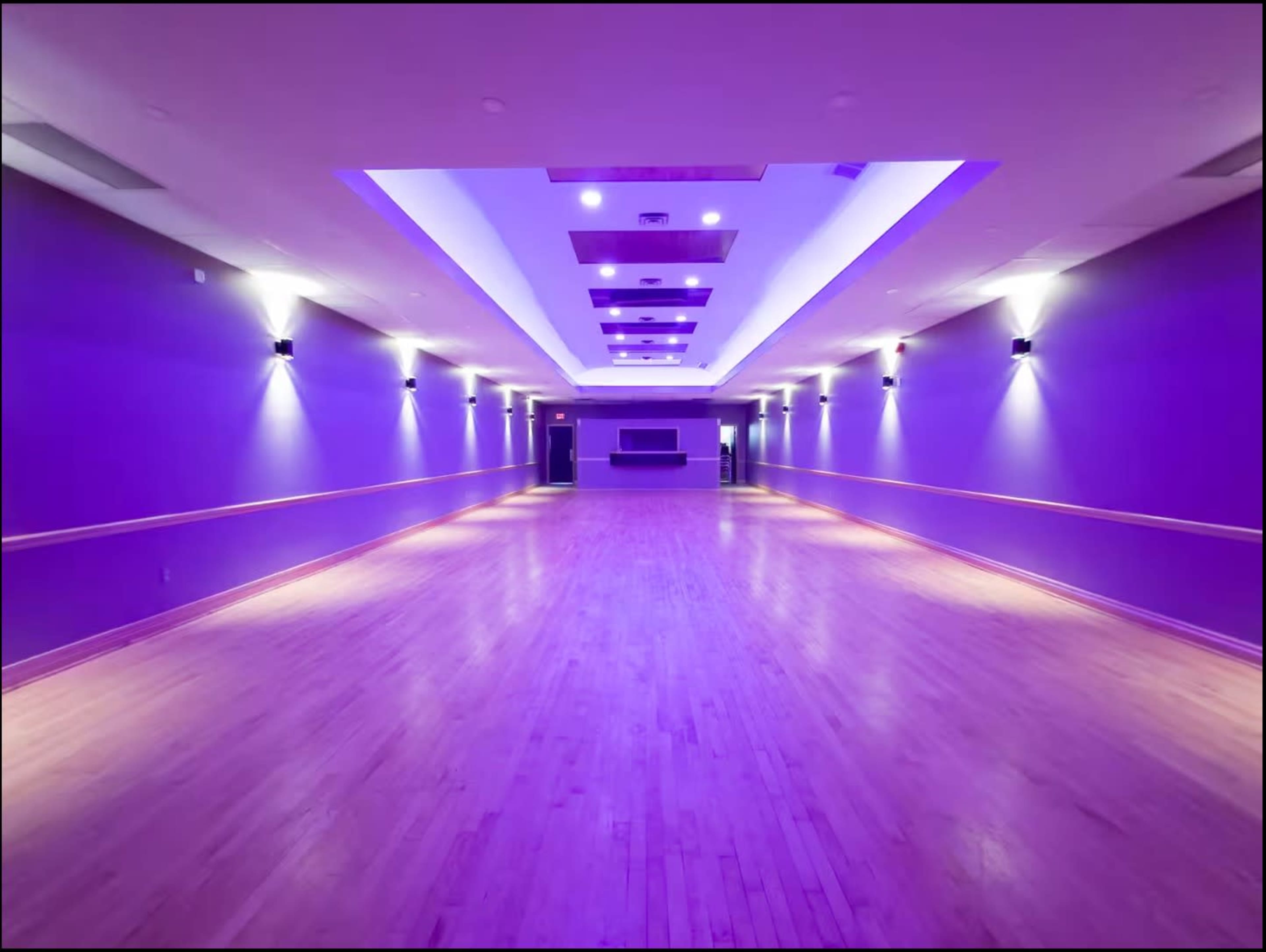A spacious dance studio with wooden flooring, illuminated by purple lights and lined with walls painted in a similar shade.