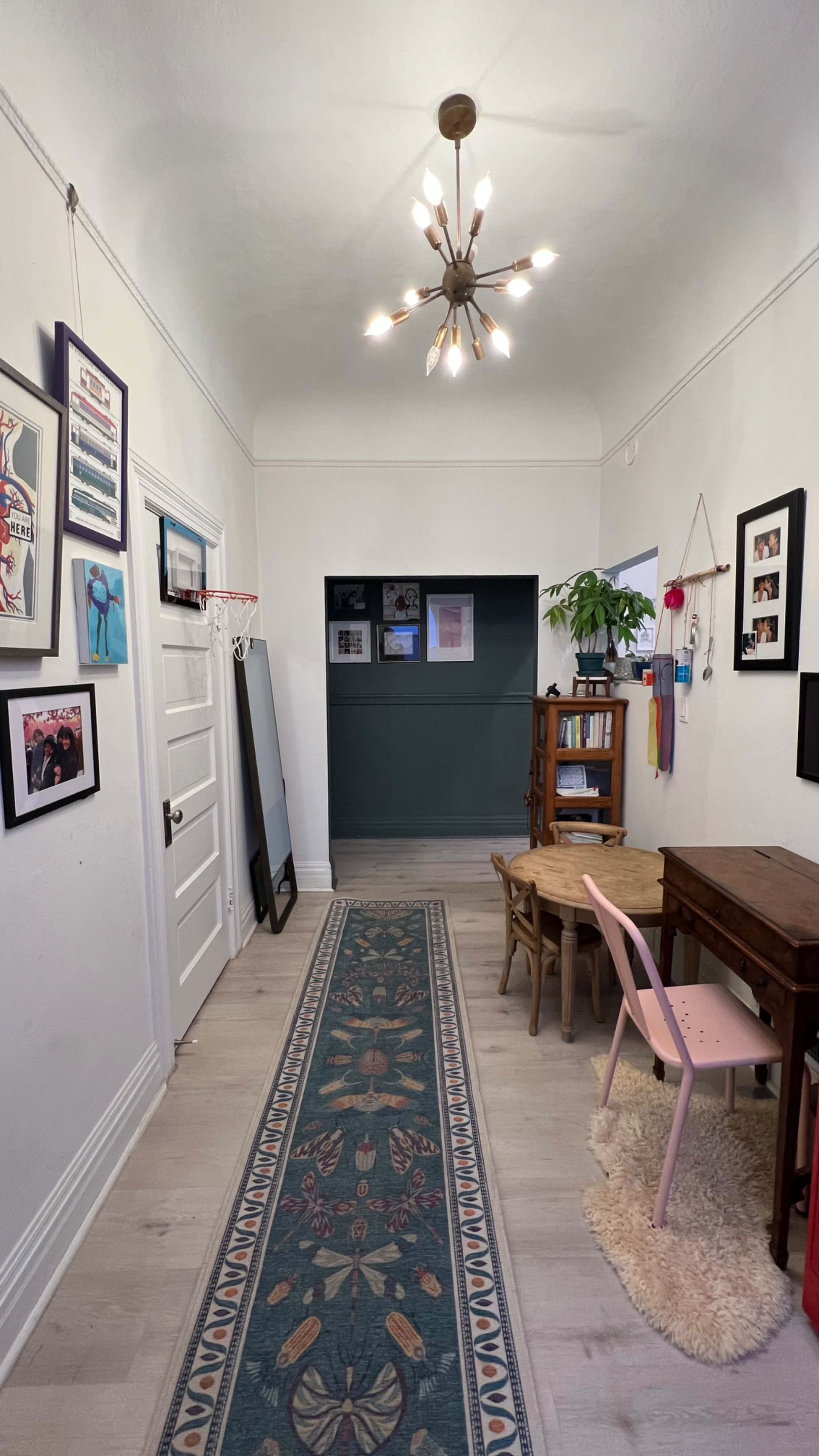 The image shows a hallway with a patterned runner rug, a chandelier, framed pictures on the walls, and furniture including a small table and chairs.