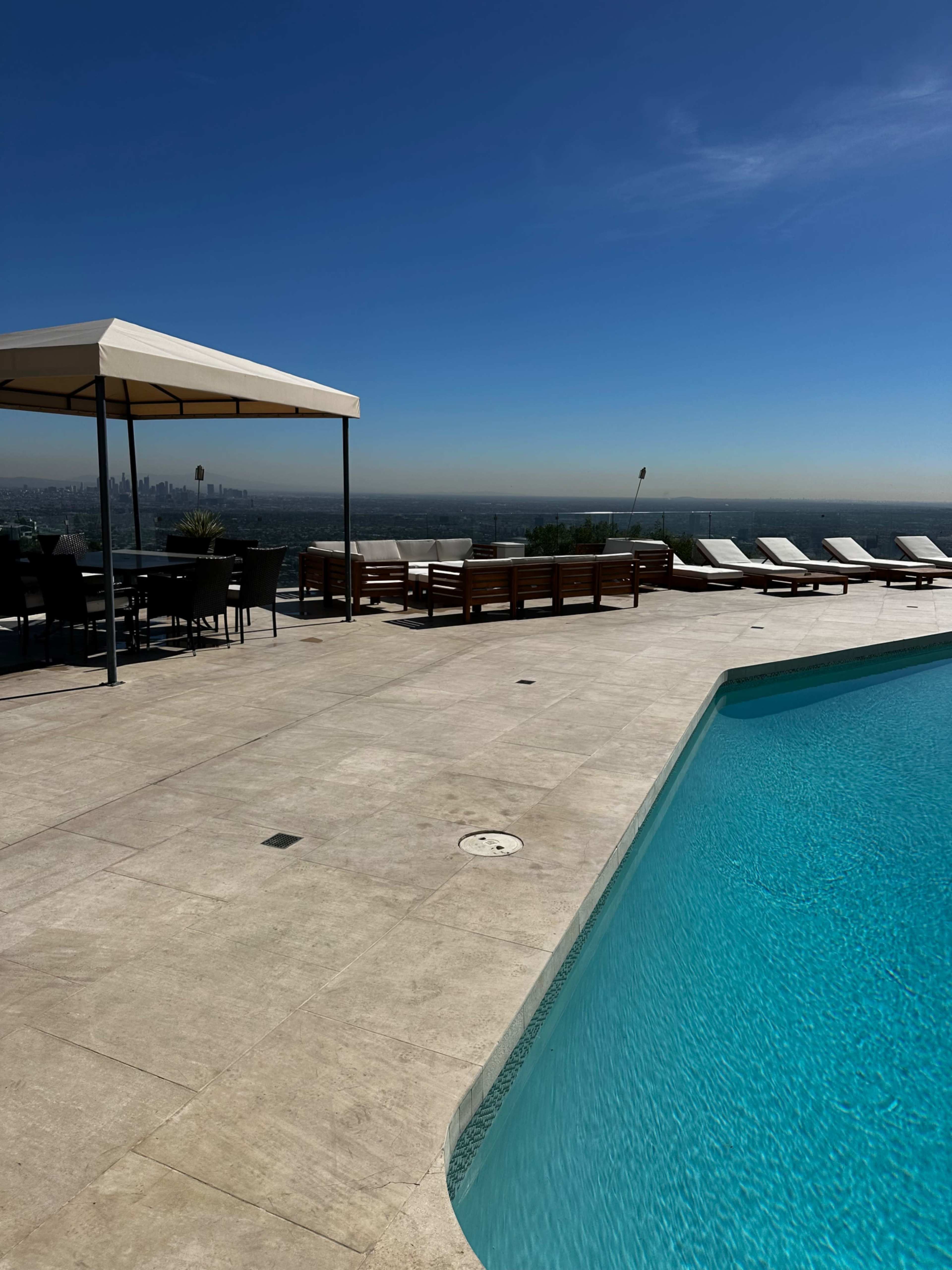 A clear blue pool is surrounded by lounge chairs and a dining area with a canopy on a sunlit terrace overlooking a cityscape.