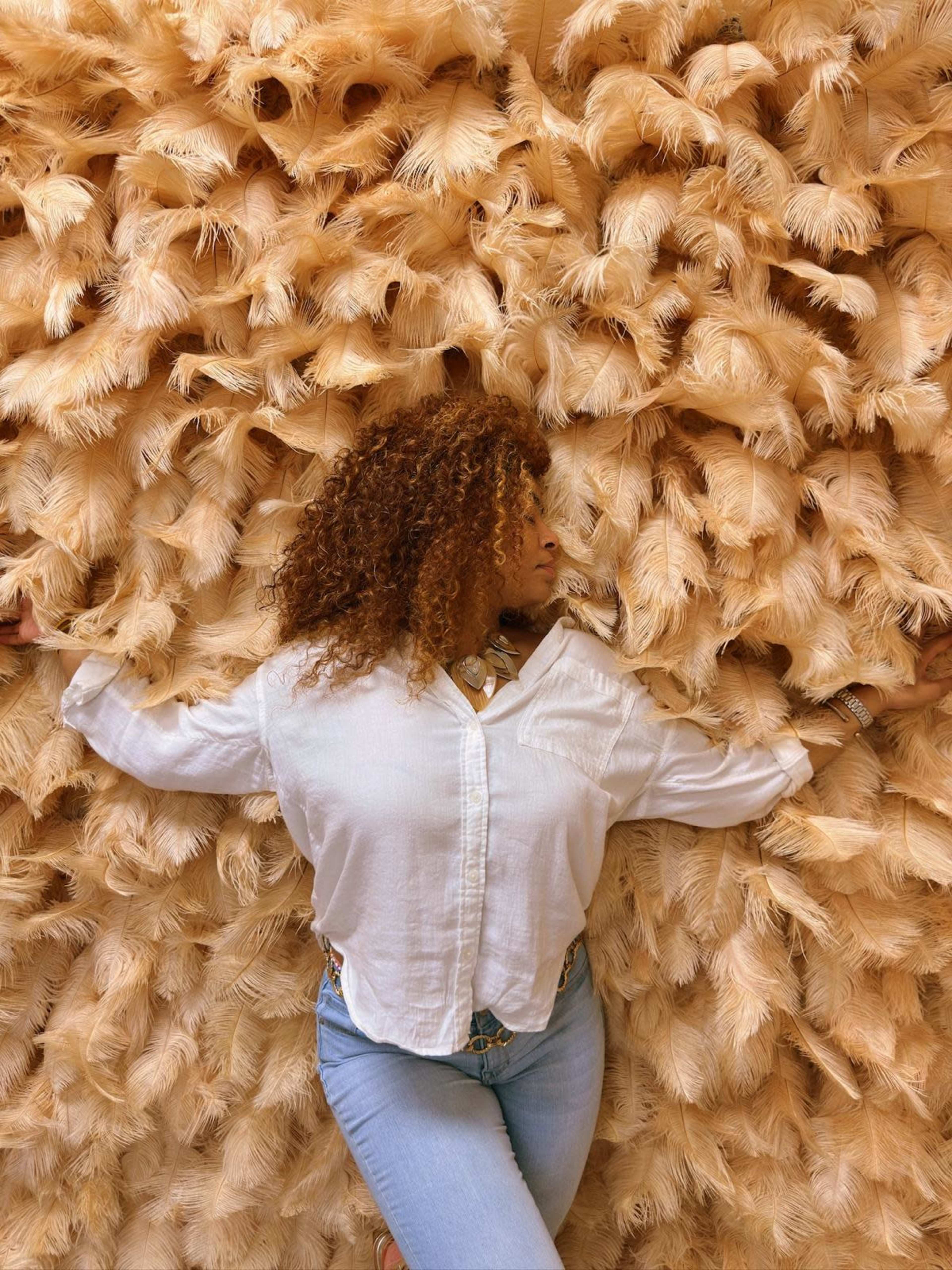 A person with curly hair is posing against a wall covered in a textured, beige material that resembles feathers.