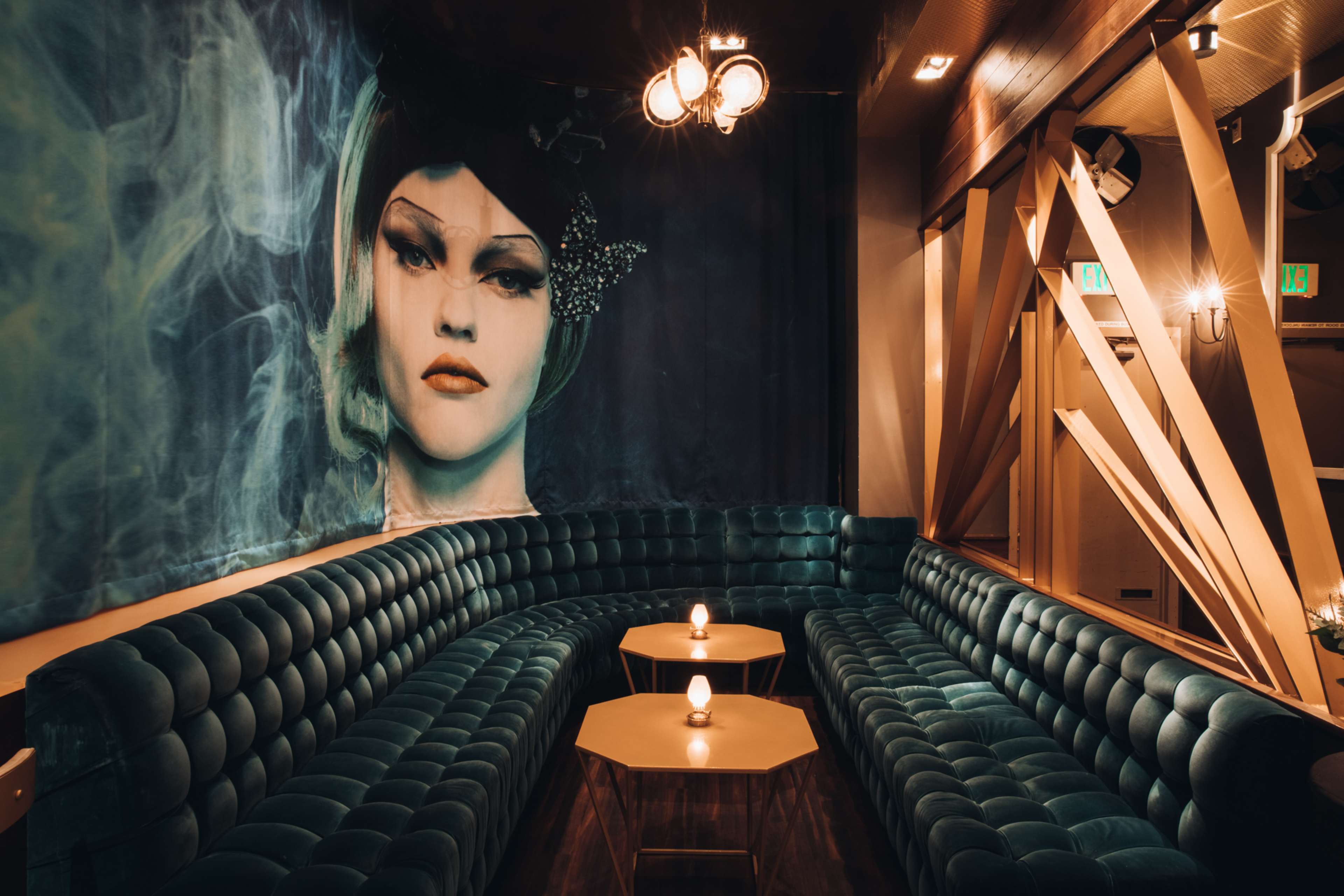 A plush, curved green velvet sofa is positioned against a wall featuring a large mural of a woman's face, with two small tables in front holding lit candles.