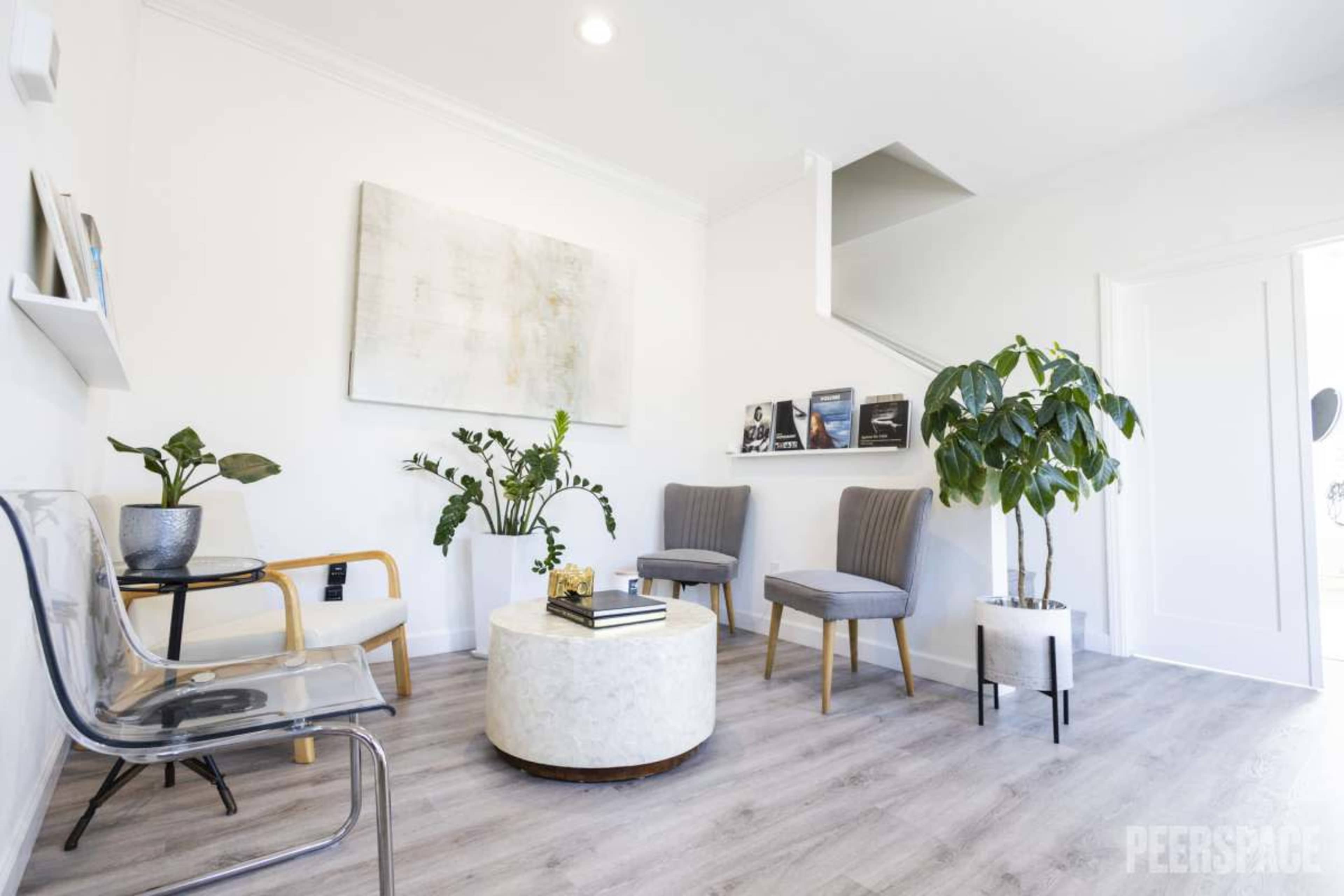 The image shows a modern waiting area with a circular coffee table, two gray chairs, a clear plastic chair, and decorative plants against a white wall.