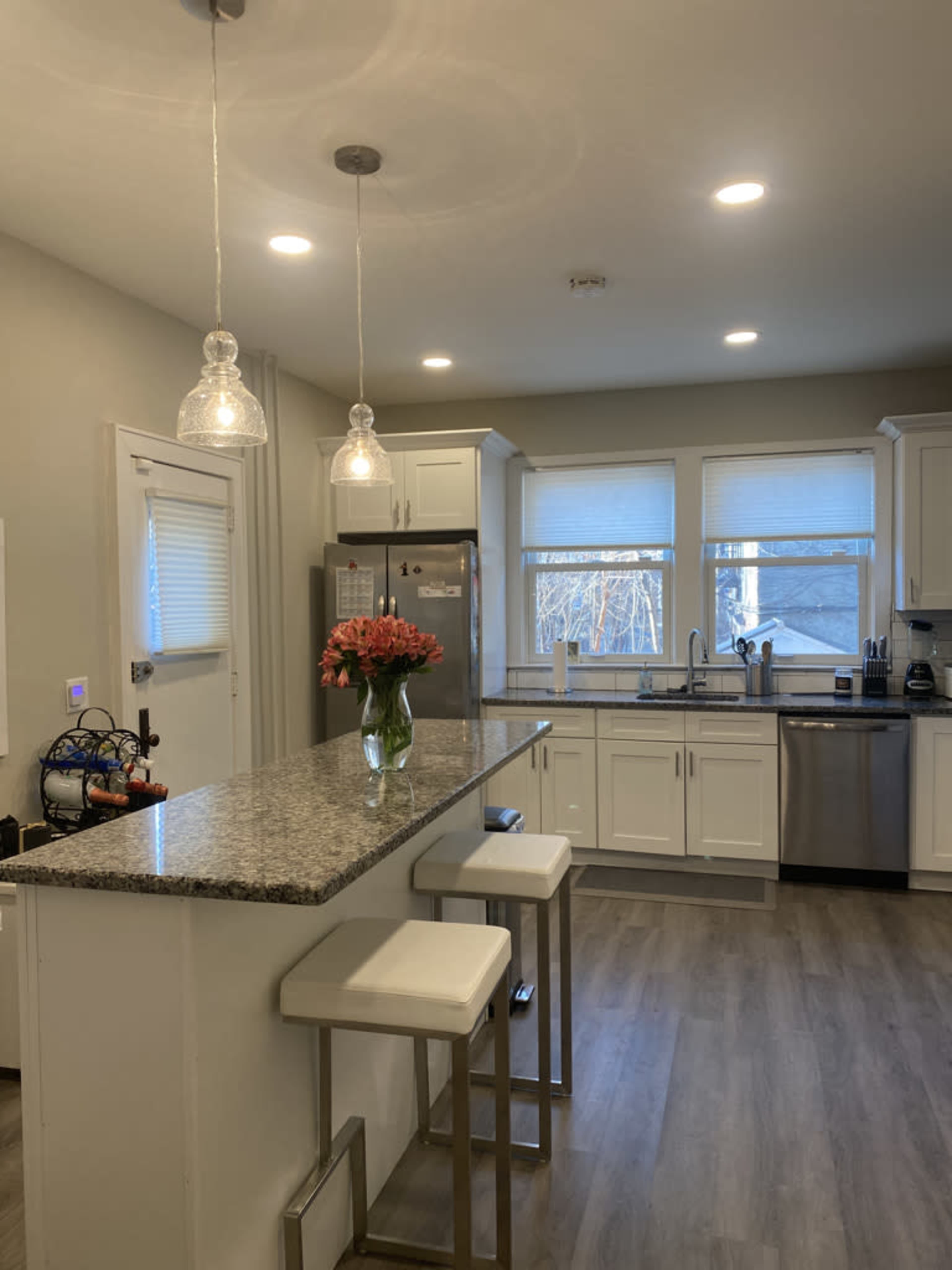 A modern kitchen features white cabinetry, a granite countertop island with seating, and stainless steel appliances, illuminated by pendant lighting.