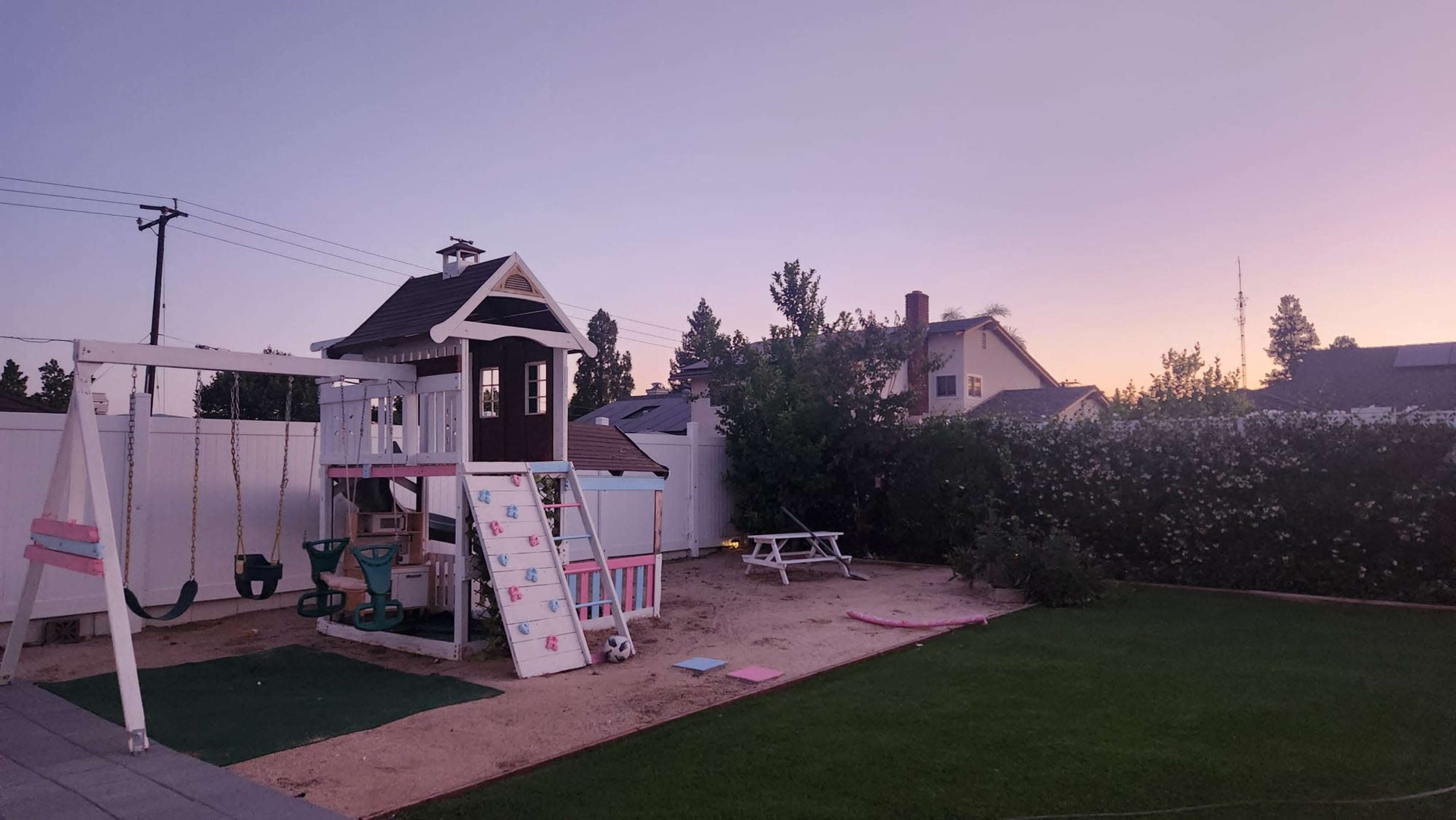 A playground with a swing set and playhouse is situated in a sandy backyard, surrounded by grass and trees at dusk.