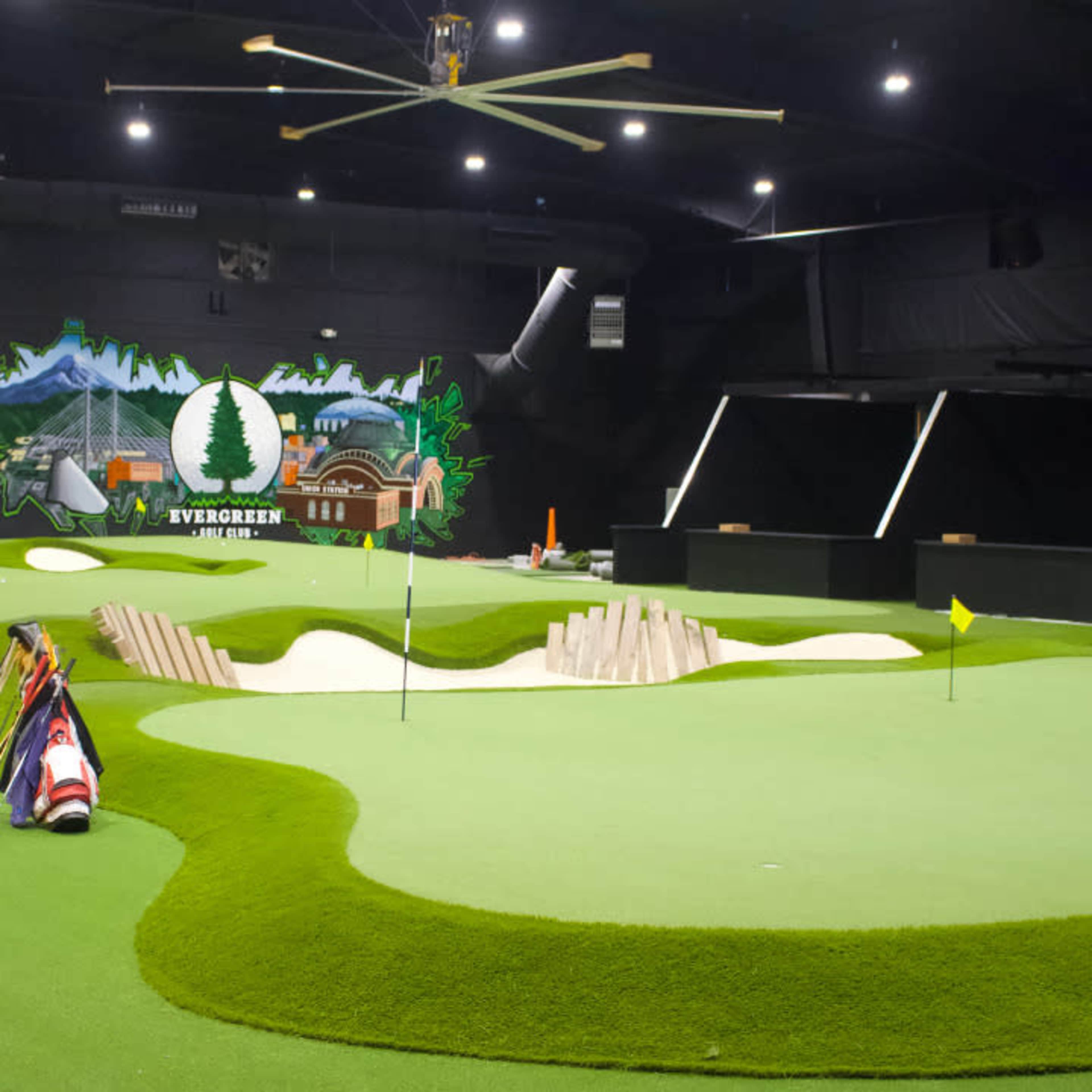 The image shows an indoor mini-golf course featuring artificial grass, a mural on the wall, and various putting greens.