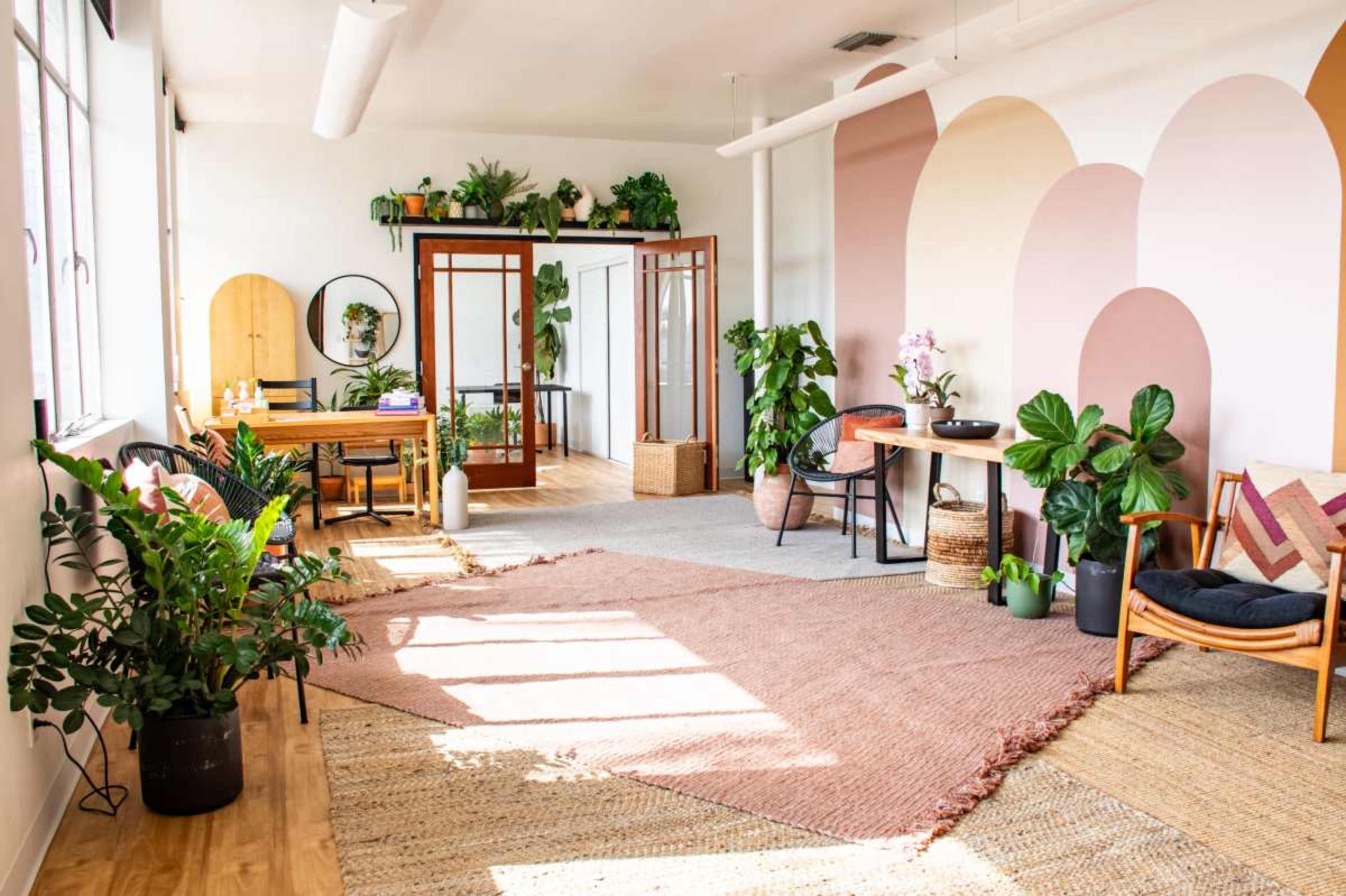 The image shows a bright, modern interior space with wooden floors, various plants, a dining area, and colorful wall designs.